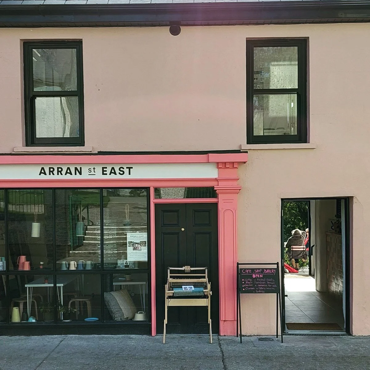 A pastel pink and black building with a sign that reads "ARRAN ST EAST." The ground floor has large windows showing inside seating and mugs. A black door is centered, flanked by pink decorative trim. To the right, an open doorway, and a chalkboard sign indicates a cafe, shop, and bakery with an open sign.