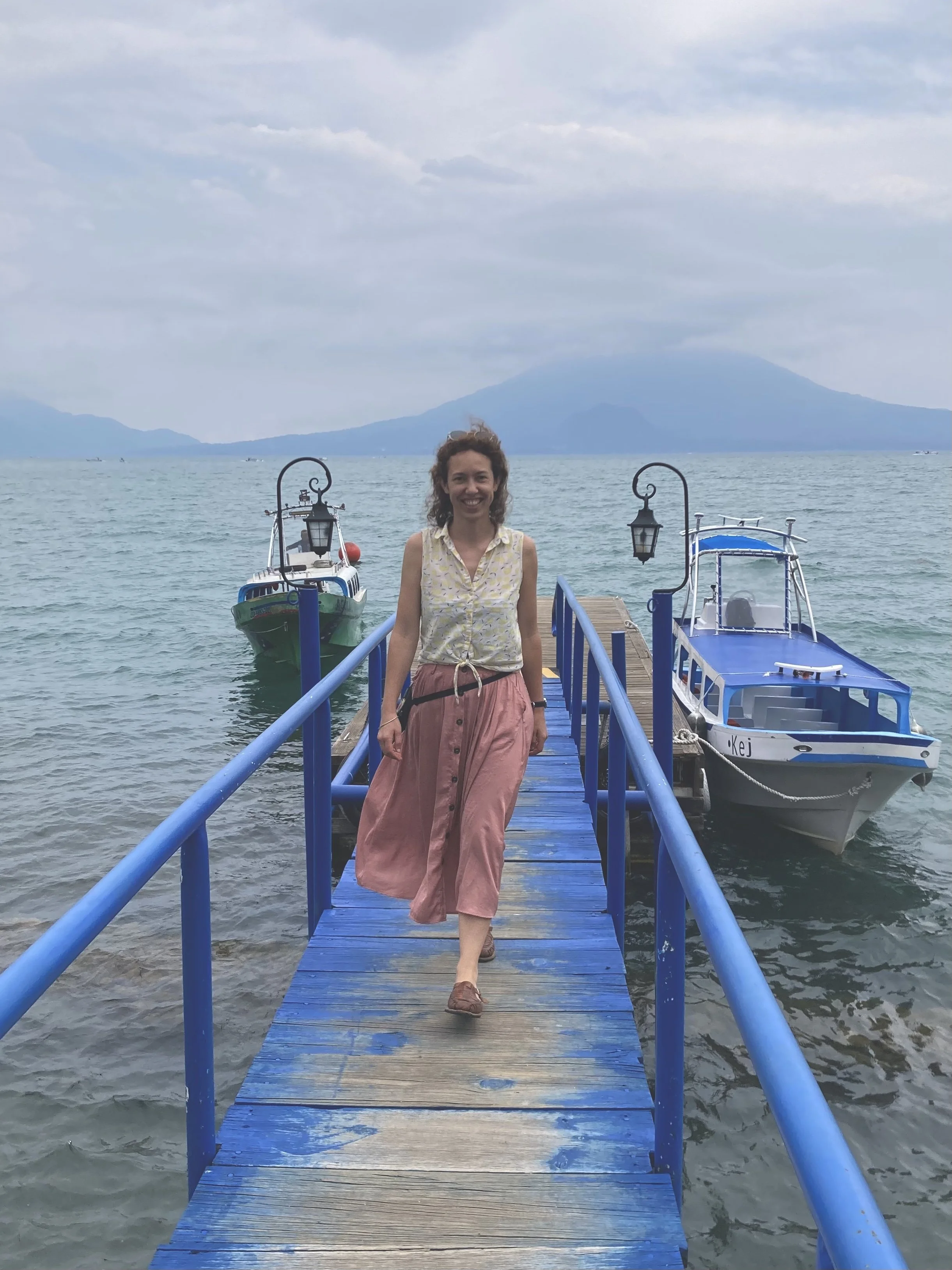 A woman walking on a blue wooden dock over water, with boats on both sides and mountains in the background, during daytime.