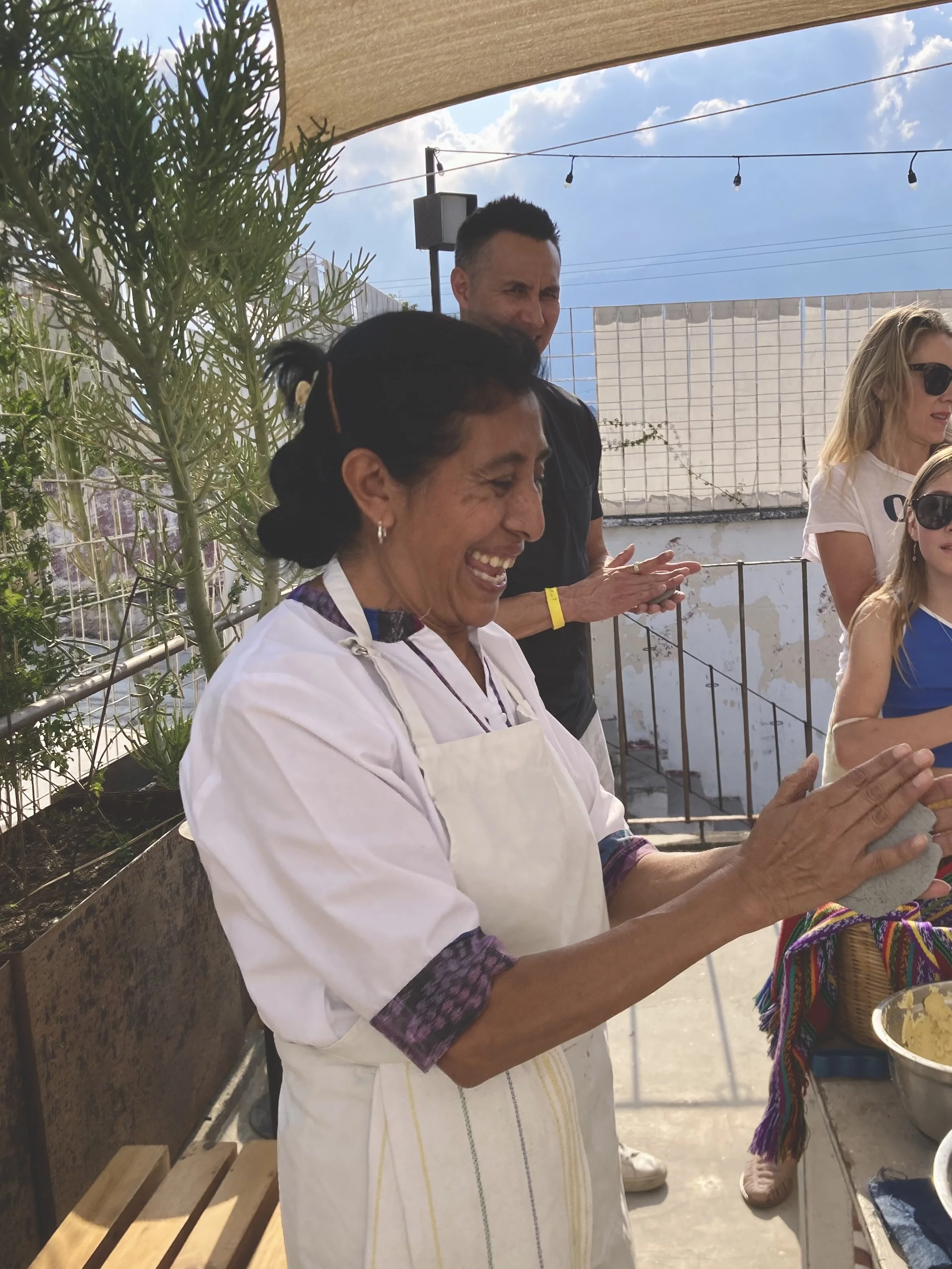 A woman dressed in a white apron is smiling while preparing food at an outdoor gathering. She is surrounded by other people, including a man in a black shirt and a woman with sunglasses, under a beige canopy with string lights. There are plants and a white fence in the background.