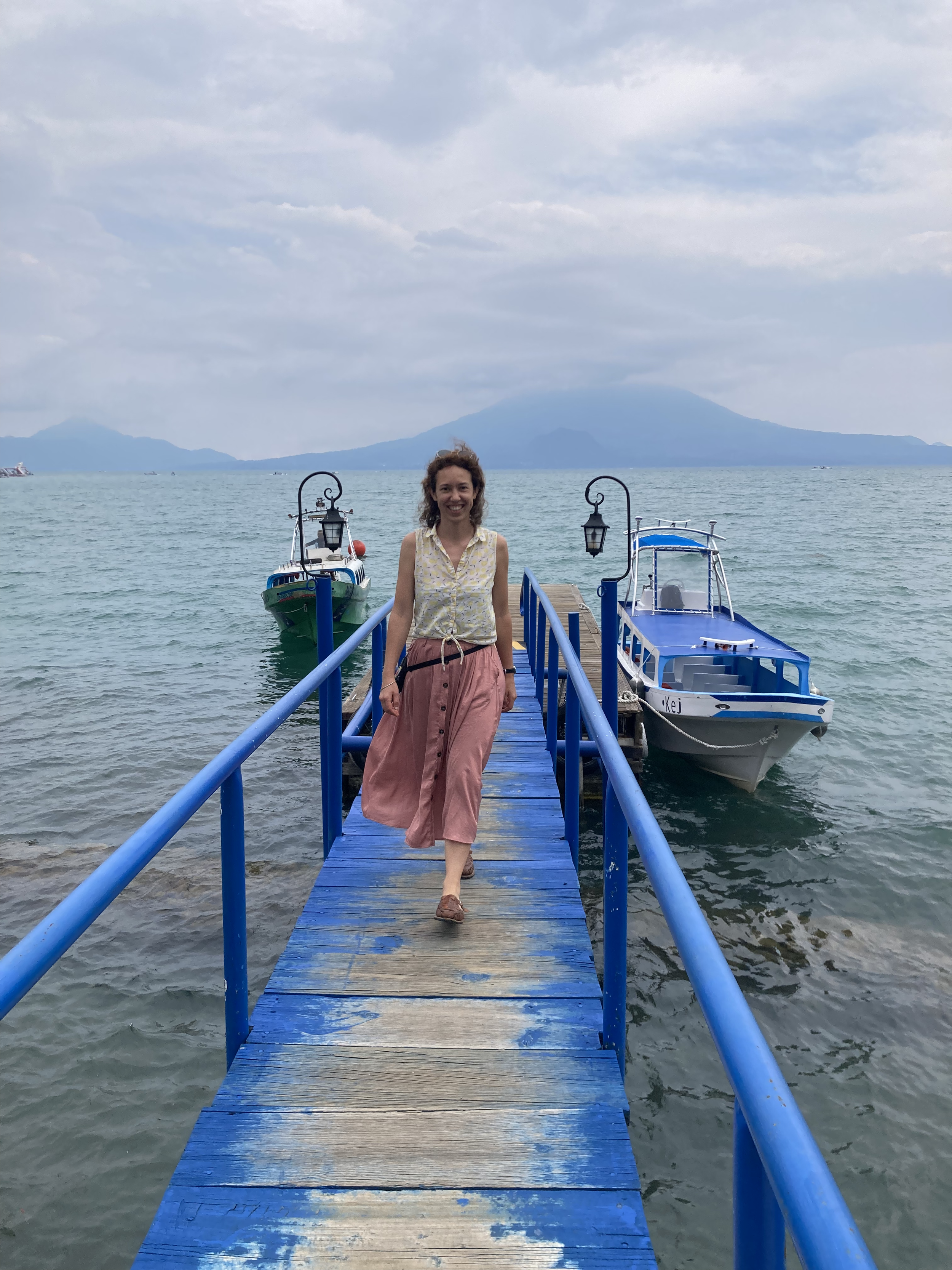 A woman walking on a blue wooden pier over water, with boats on the water and mountains in the background.