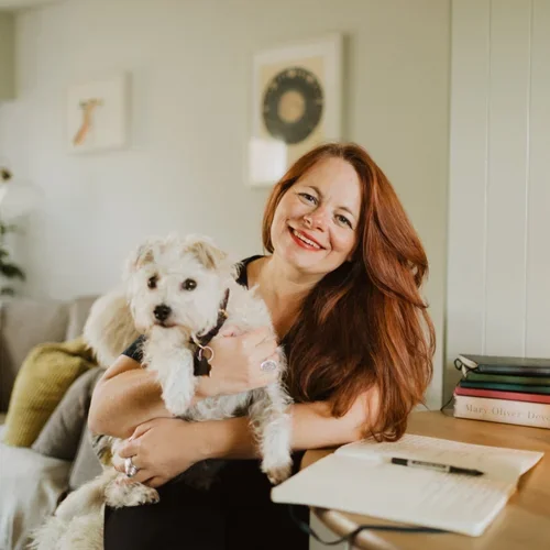 A woman with long red hair smiling and holding a small white dog inside a home.