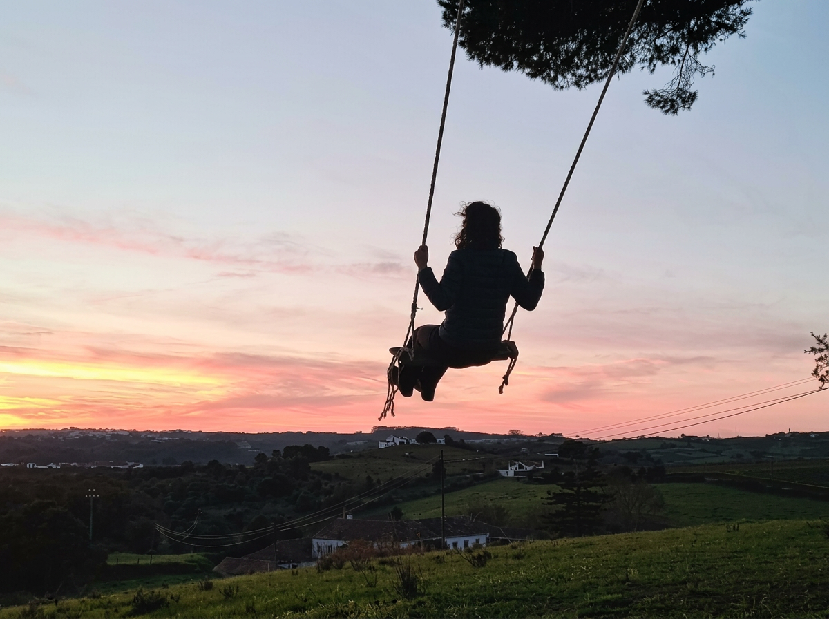 Person on a swing outdoors at sunset over a rural landscape with houses and trees in Portugal.