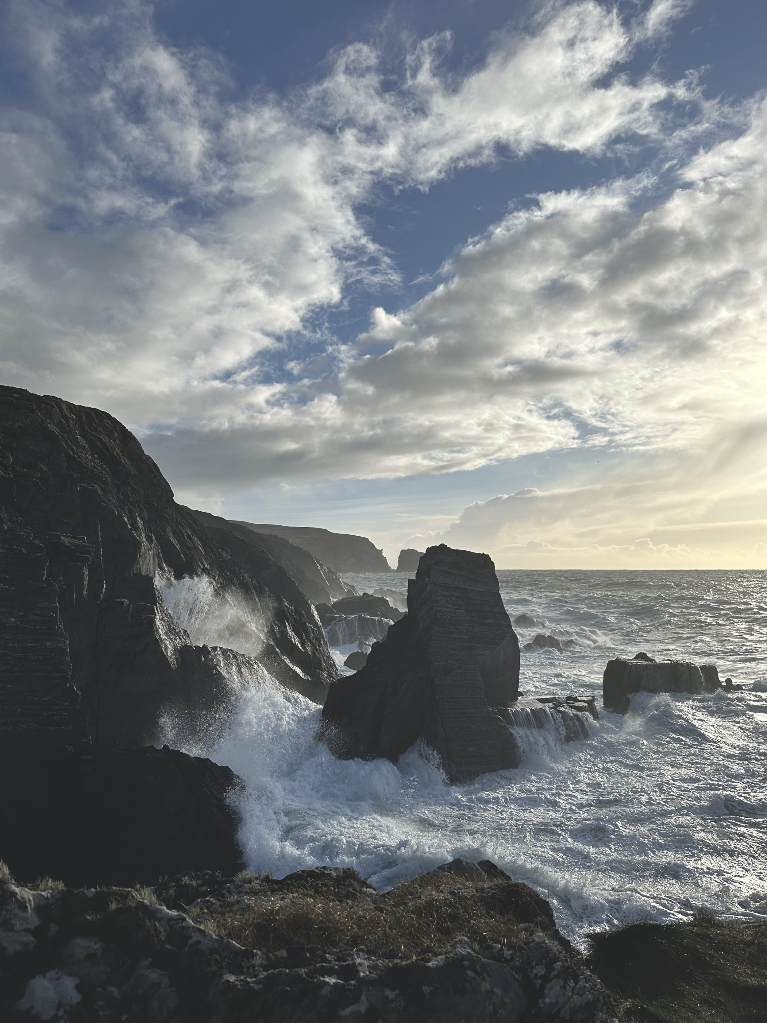 Coastal cliffs with crashing waves, cloudy sky with patches of blue, and rocky shoreline at sunset.