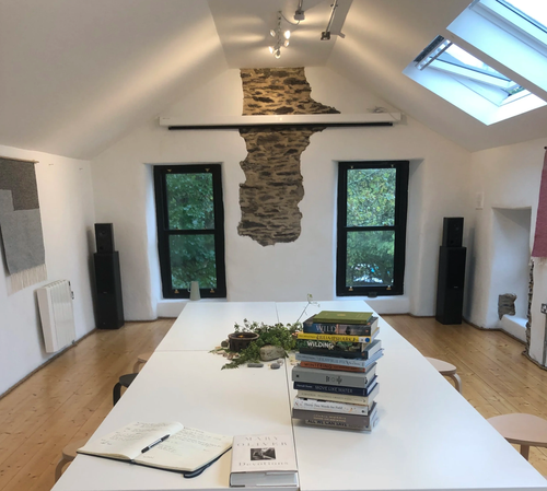 Room with a white table, open notebook, plants, and a stack of books. Two black speakers, two windows, exposed brick wall, and skylight in the ceiling.