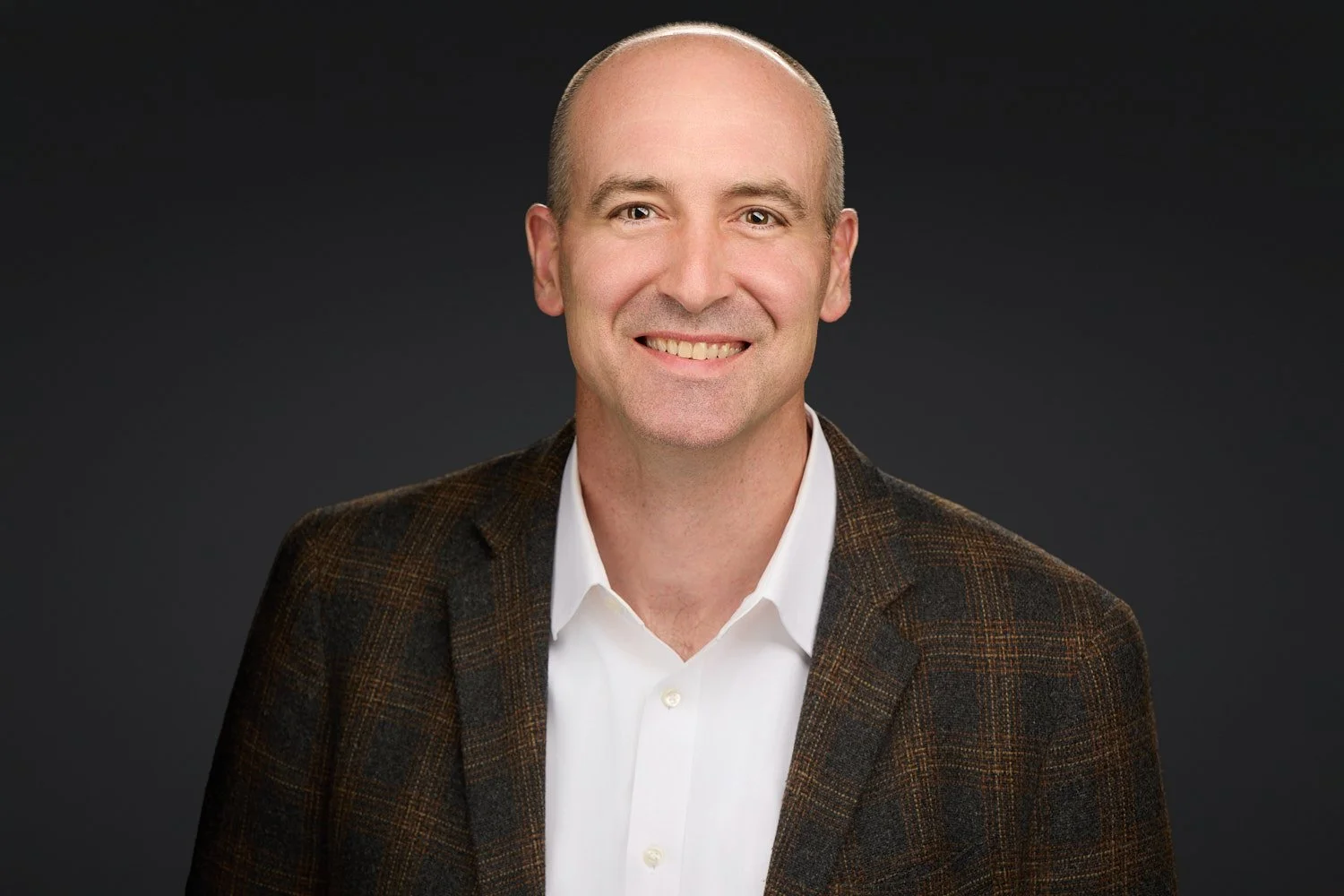 A smiling, bald man in a white shirt and brown plaid blazer poses against a dark background.