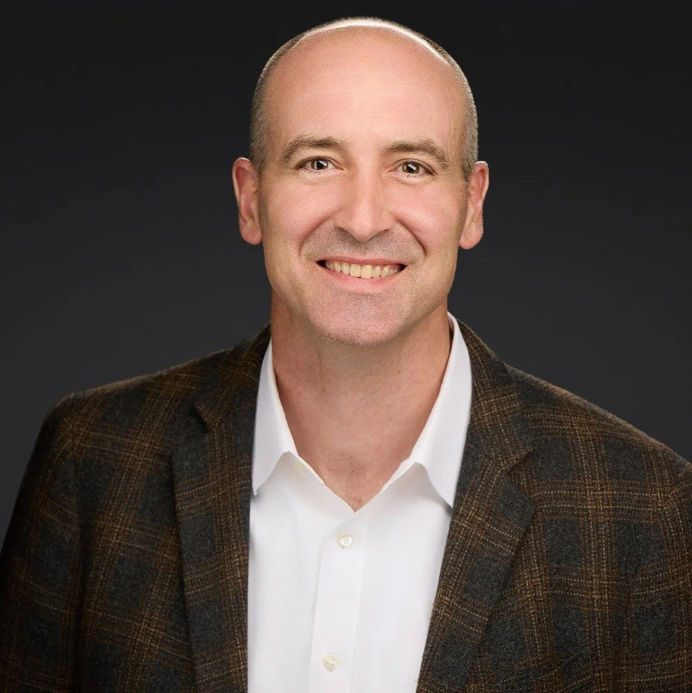 Headshot of a smiling bald man in a white shirt and brown blazer against a dark background.