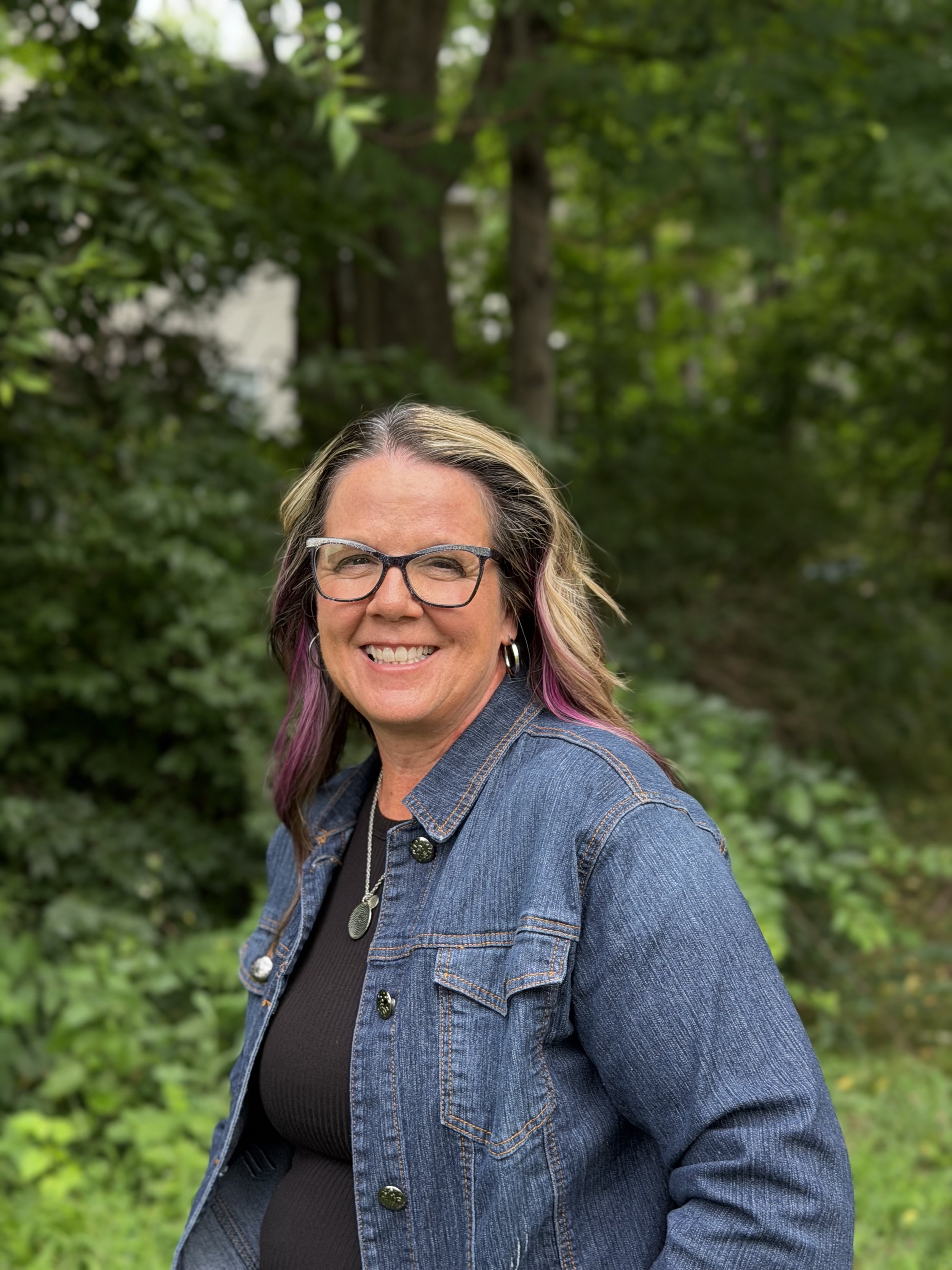 A woman smiling outdoors in front of green trees, wearing glasses, a denim jacket, and a black top.