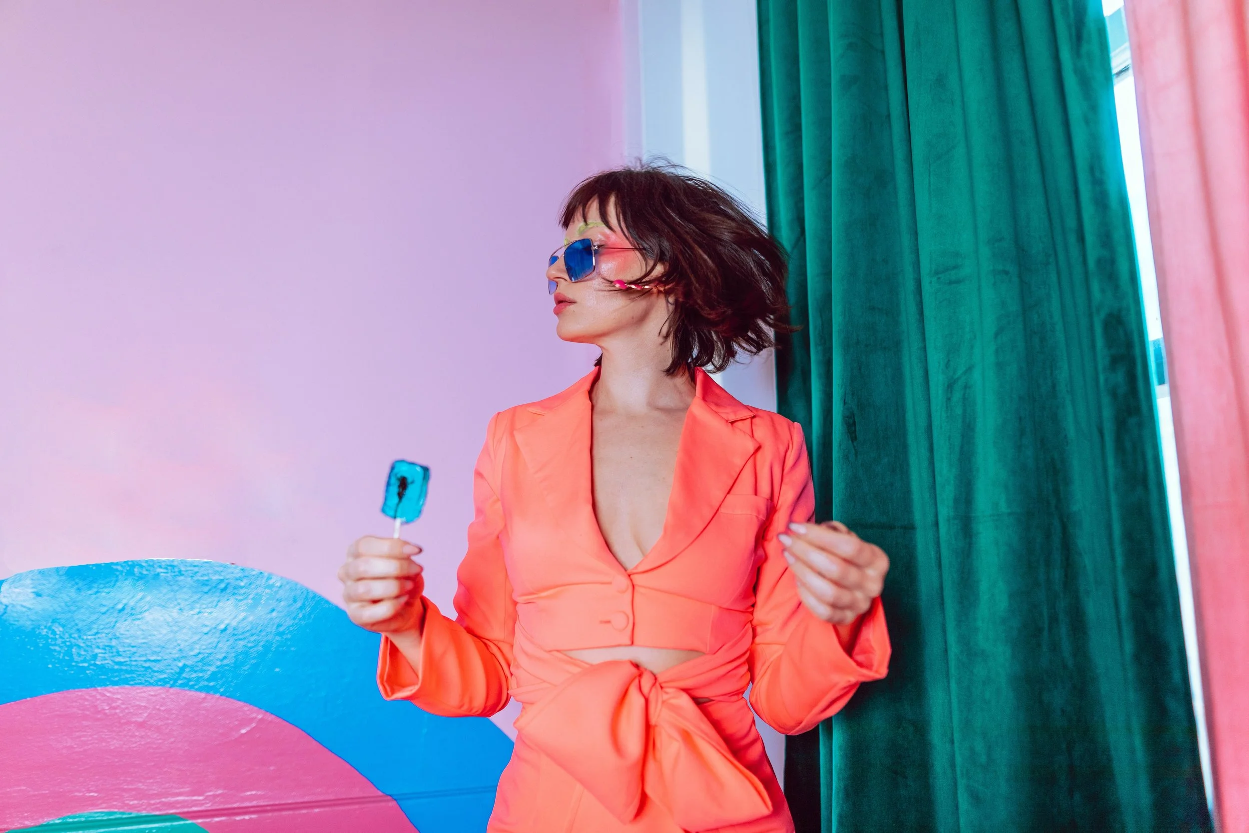 Woman with short dark hair wearing sunglasses and a pink blouse, holding a blue ice pop, standing in front of a colorful background with pink, blue, and green curtains.