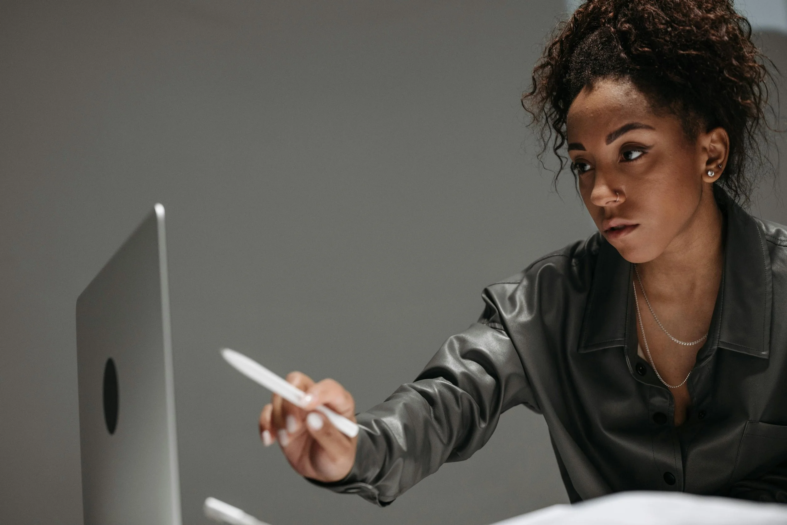 A woman with curly hair and earrings sitting at a desk, holding a pen and looking at a computer screen in a gray room.