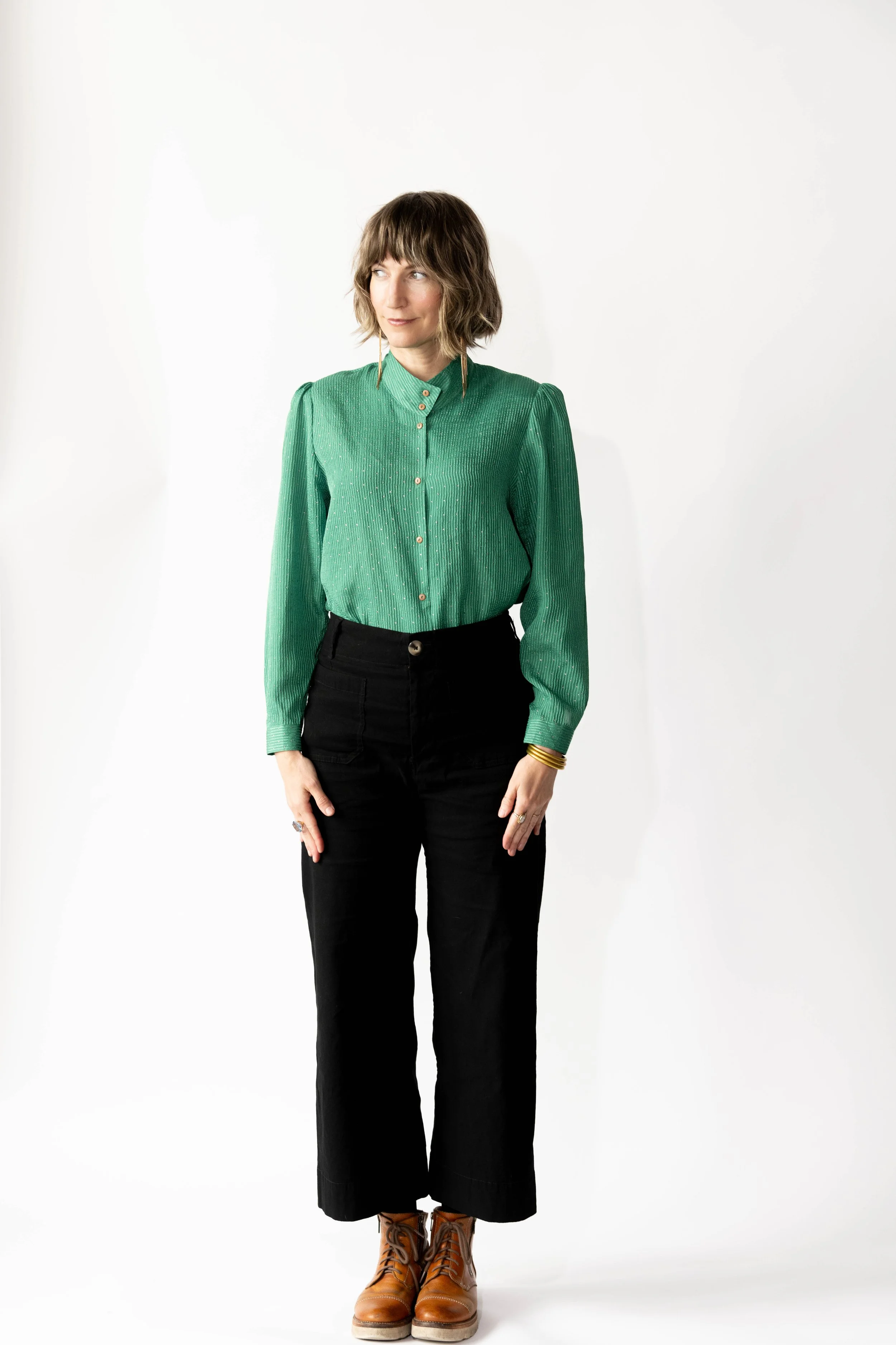 A woman with shoulder-length brown hair, wearing a green long-sleeve blouse, black pants, and brown work boots, standing against a white background. She is looking slightly to her left.
