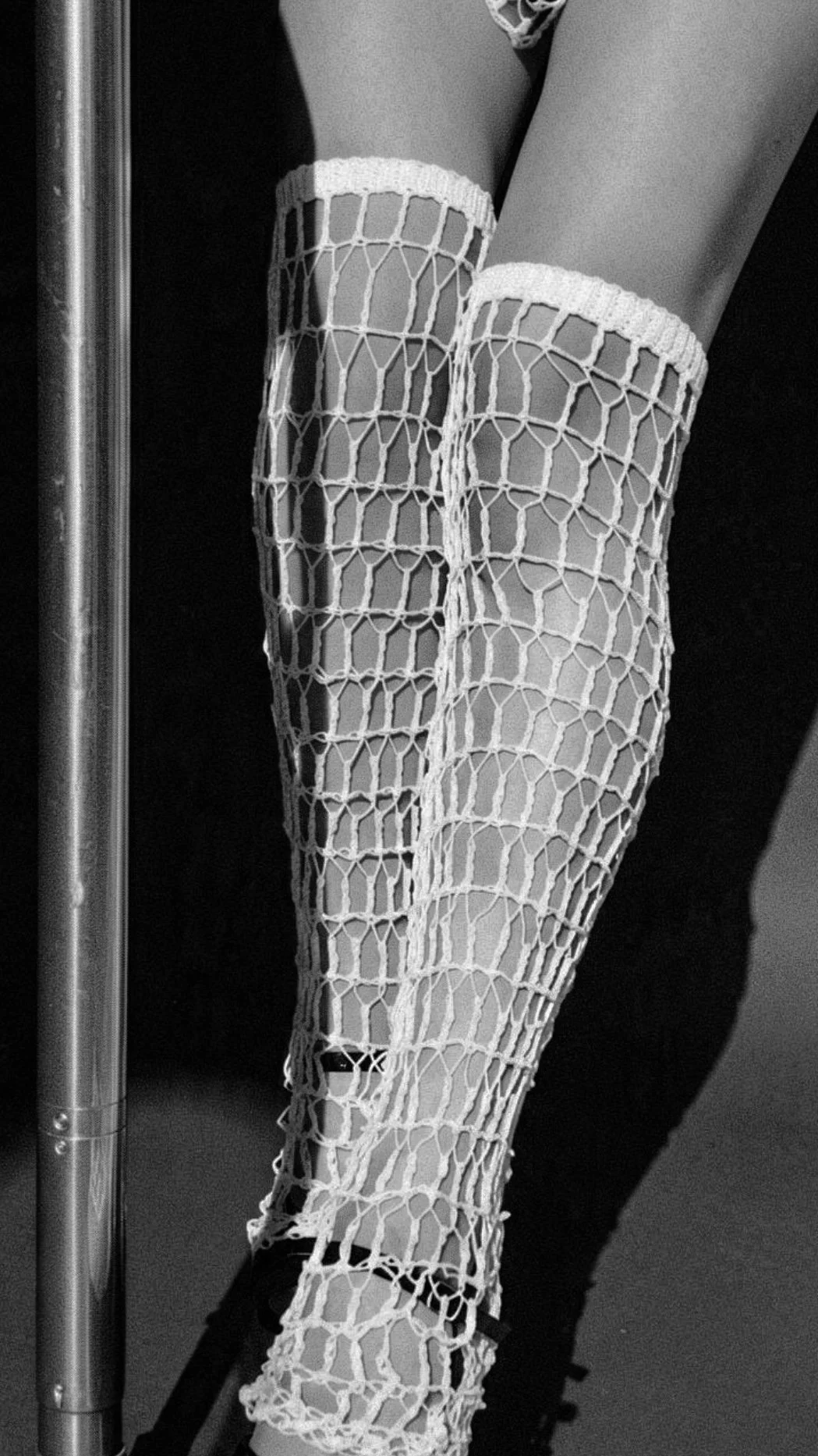 Close-up of a person's legs and ankles wrapped in white crochet mesh stockings with a grid pattern, with a metal pole and a dark floor nearby.