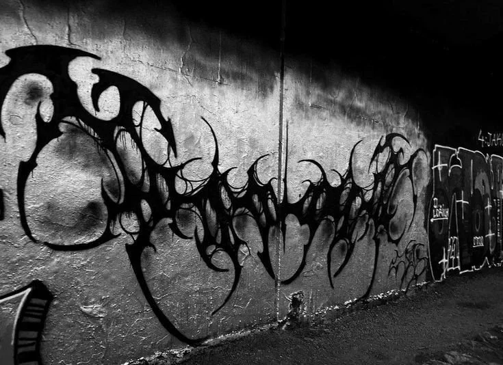 Black graffiti art on a concrete wall in an alleyway, with intricate, sharp-edged lettering and additional tags on the right side.