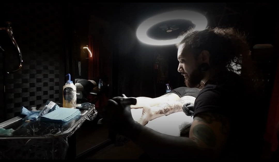 Tattoo artist working on a client's leg in a dimly lit tattoo studio, with tattoo supplies on a medical tray nearby and a ring light above.