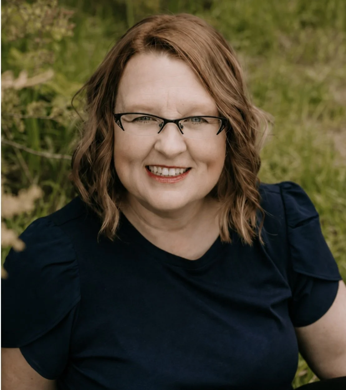 A smiling woman with shoulder-length wavy red hair and glasses, wearing a dark top, outdoors with green foliage in the background.