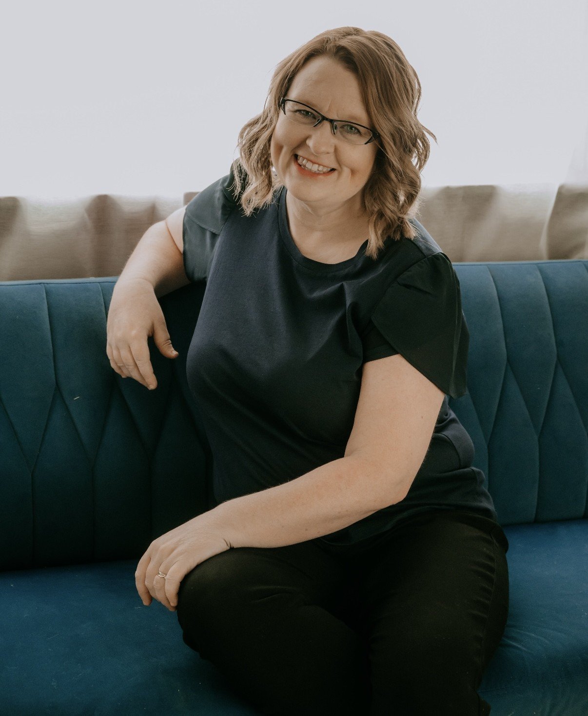 Woman with glasses and wavy, shoulder-length hair sitting on a blue sofa, smiling and leaning slightly to her right.