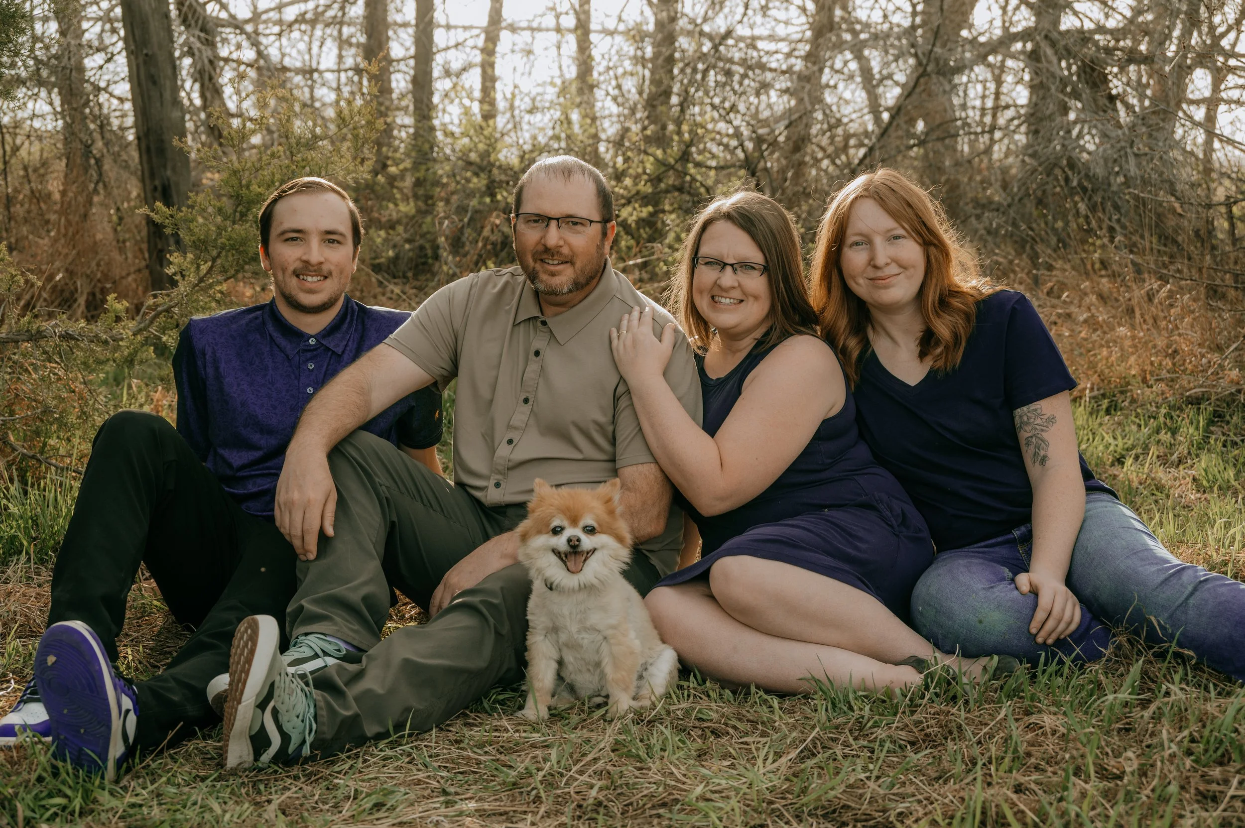 Family sitting outdoors in a wooded area with a small fluffy dog in front of them. The group includes two men and two women, all smiling, with trees and dried grass in the background.