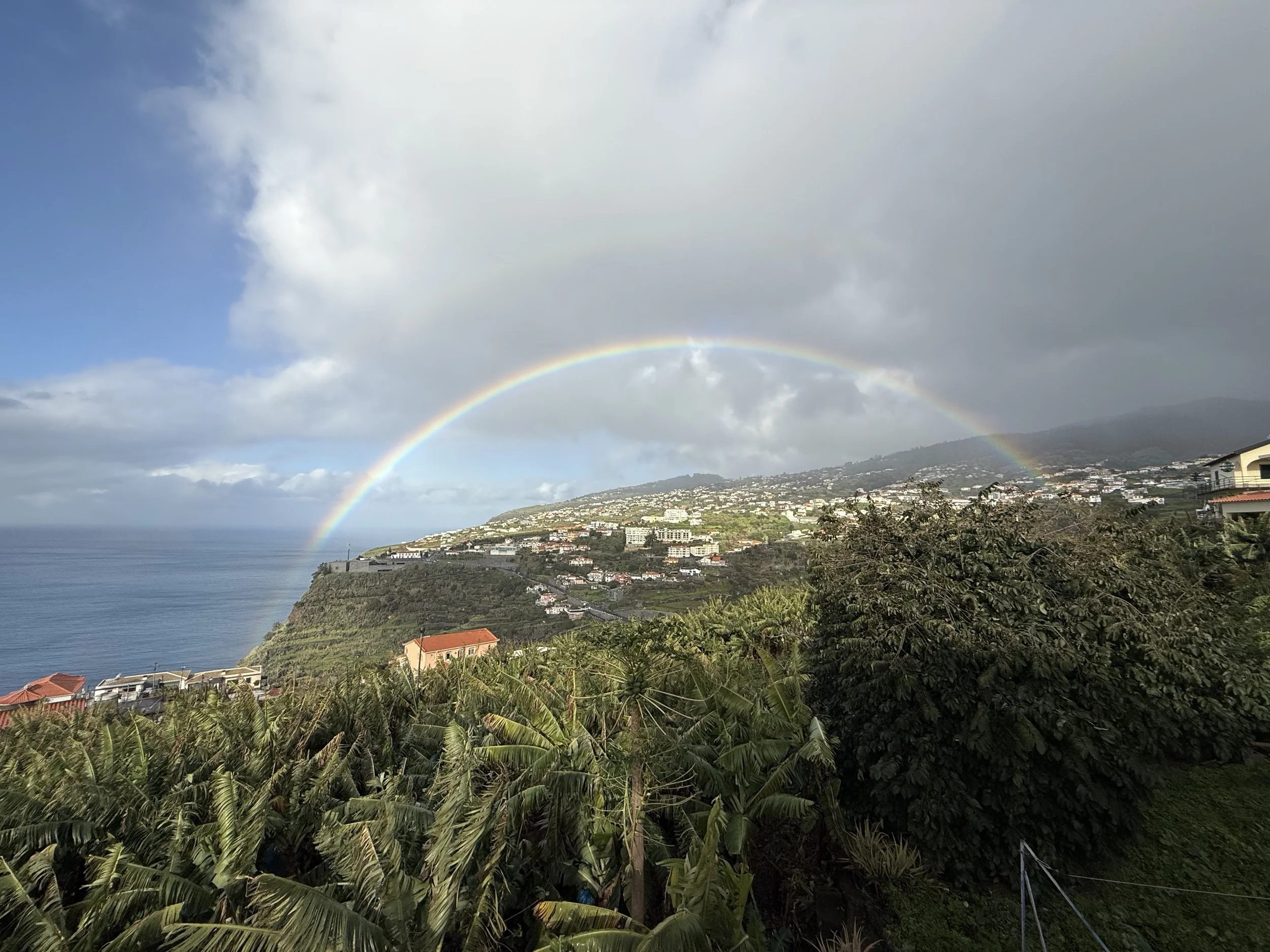 A hillside town with houses, lush green vegetation, and a double rainbow in the sky over the ocean and mountains.