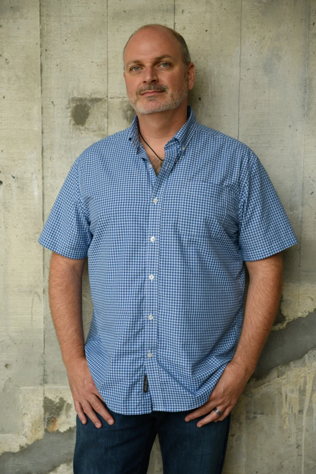 A man standing in front of a concrete wall, wearing a blue and white checkered shirt and jeans, with his hands resting on his hips.