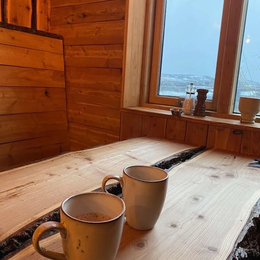Two beige mugs on a light-colored wooden table in a cabin with wooden walls and a window showing a snowy landscape outside.