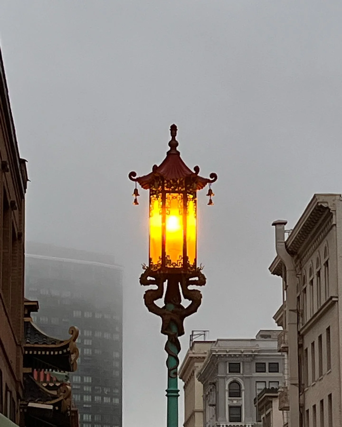 An ornate street lamp with a glowing yellow light, set against a foggy cityscape with historic buildings on either side and tall modern buildings in the background.