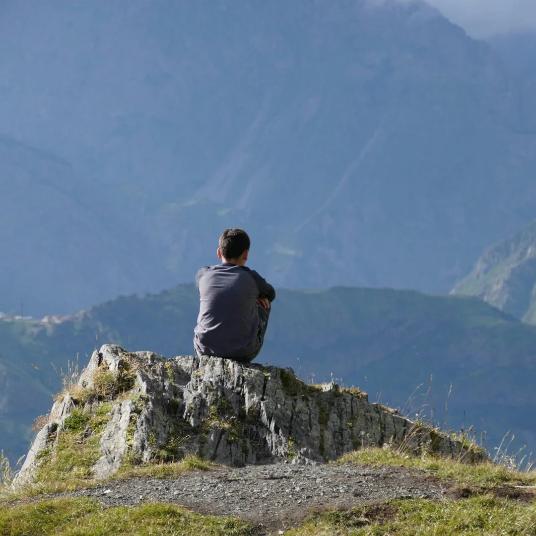 A person sitting on a rocky outcrop overlooking a mountain landscape.