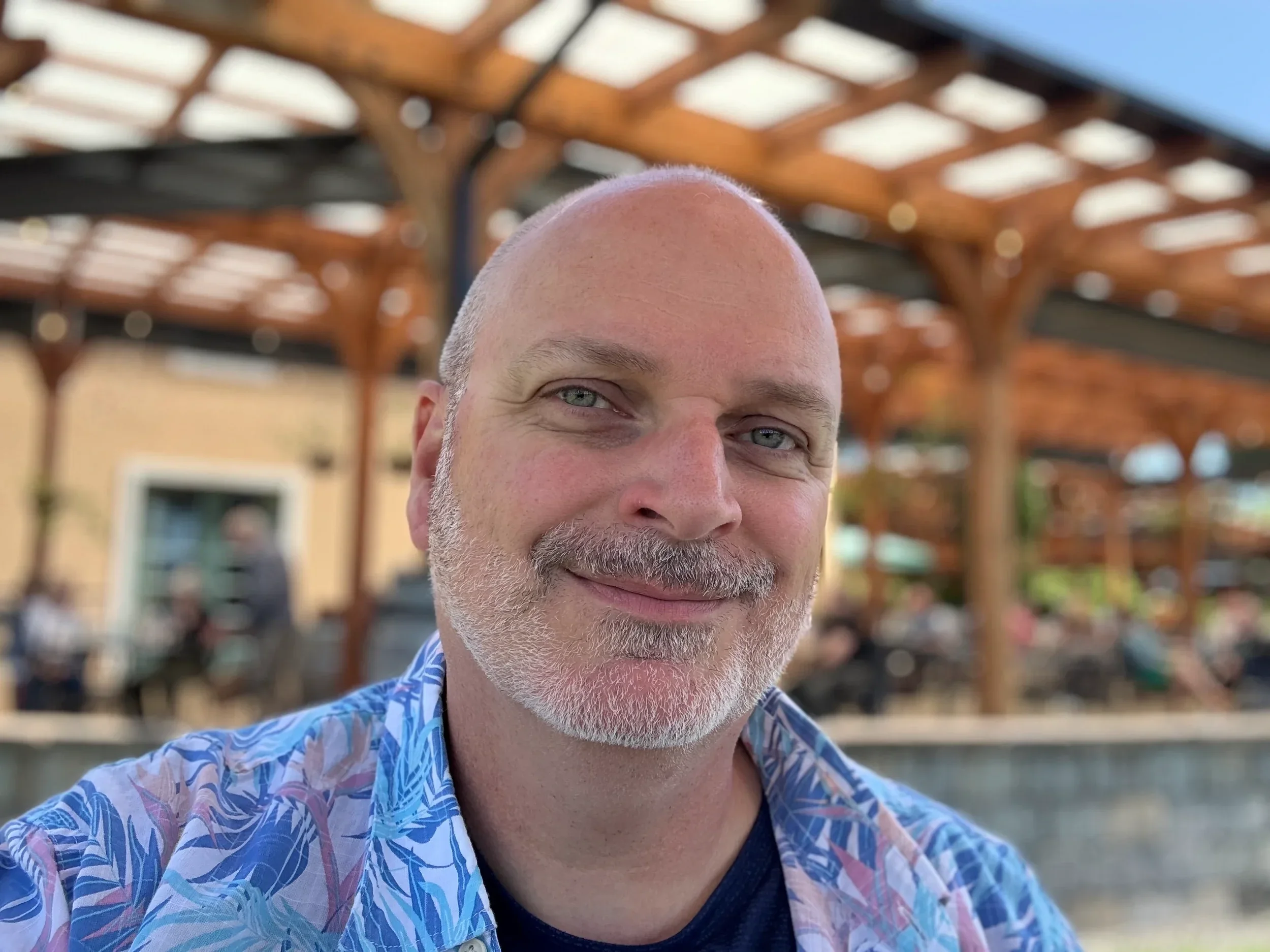 A middle-aged man with a bald head and white beard smiling outdoors, wearing a colorful patterned shirt, with a background of wooden architectural structure and a building.