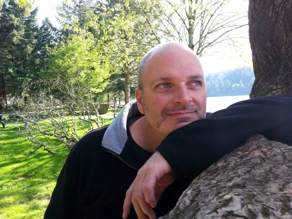 A man resting his head on a tree trunk outdoors, with green grass, trees, and a lake in the background.