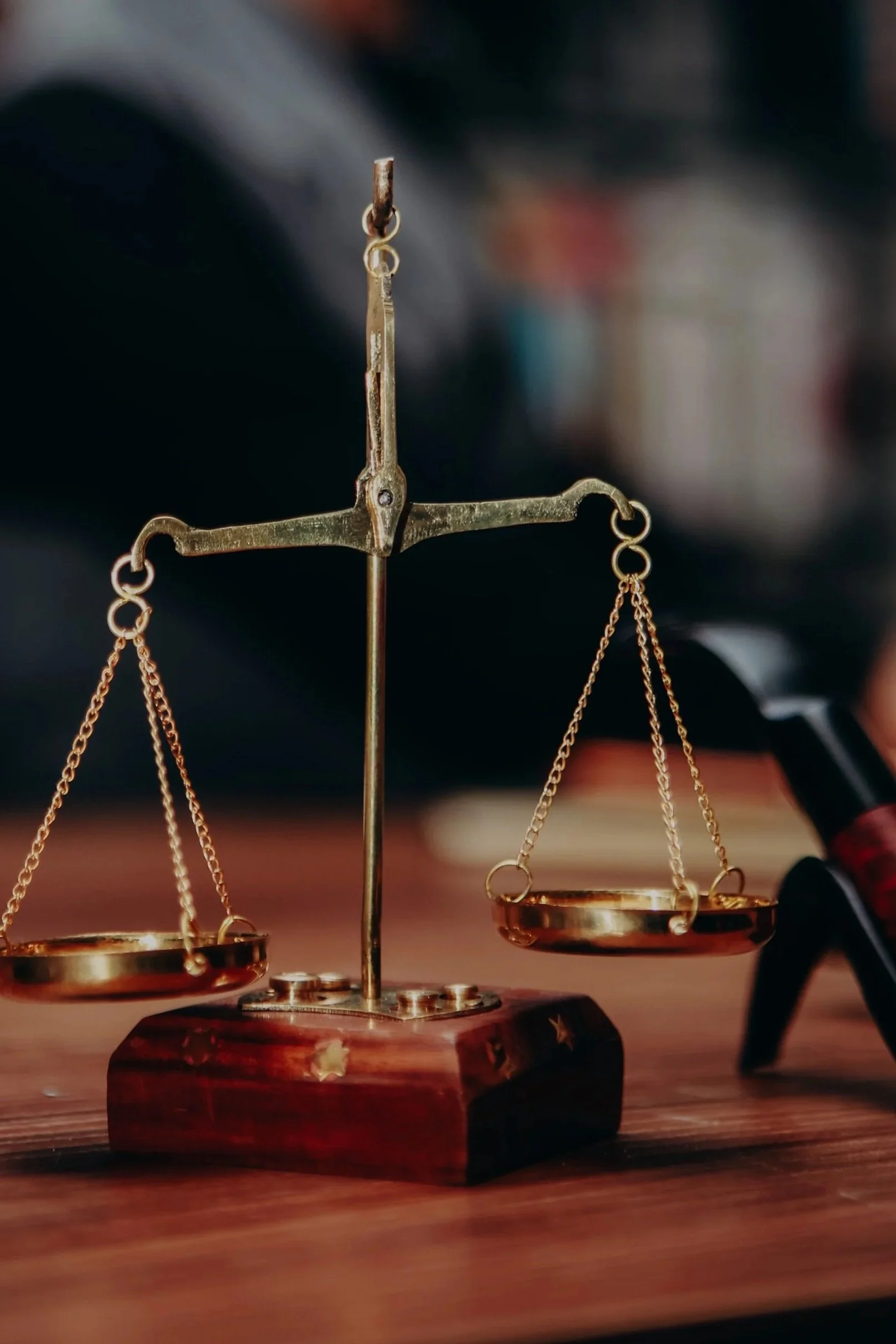 A close-up of a small gold balance scale with two pans, sitting on a wooden base, with a blurred background.