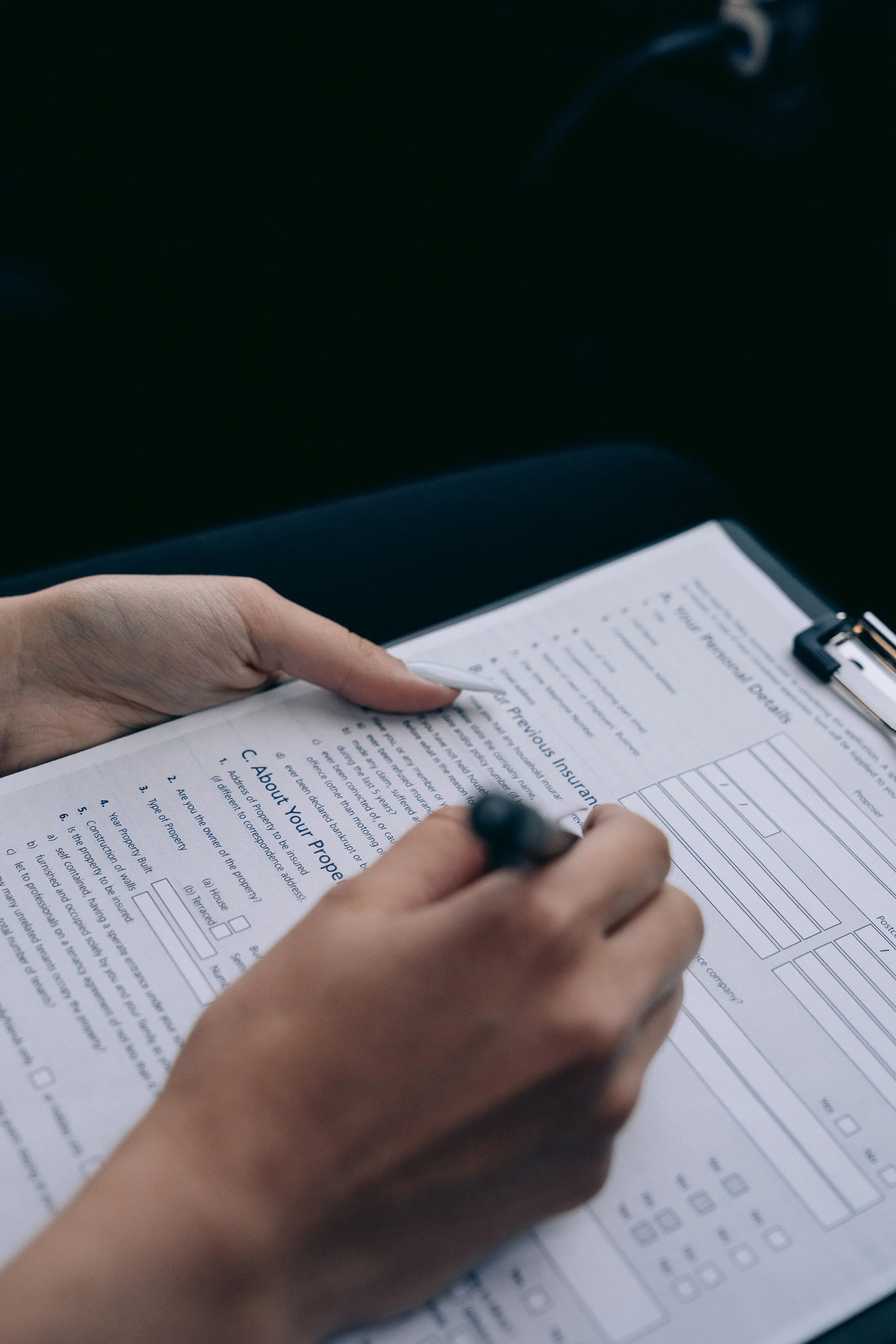 Close-up of a person's hands filling out a form on a clipboard with a pen. The form appears to be related to insurance or property details.