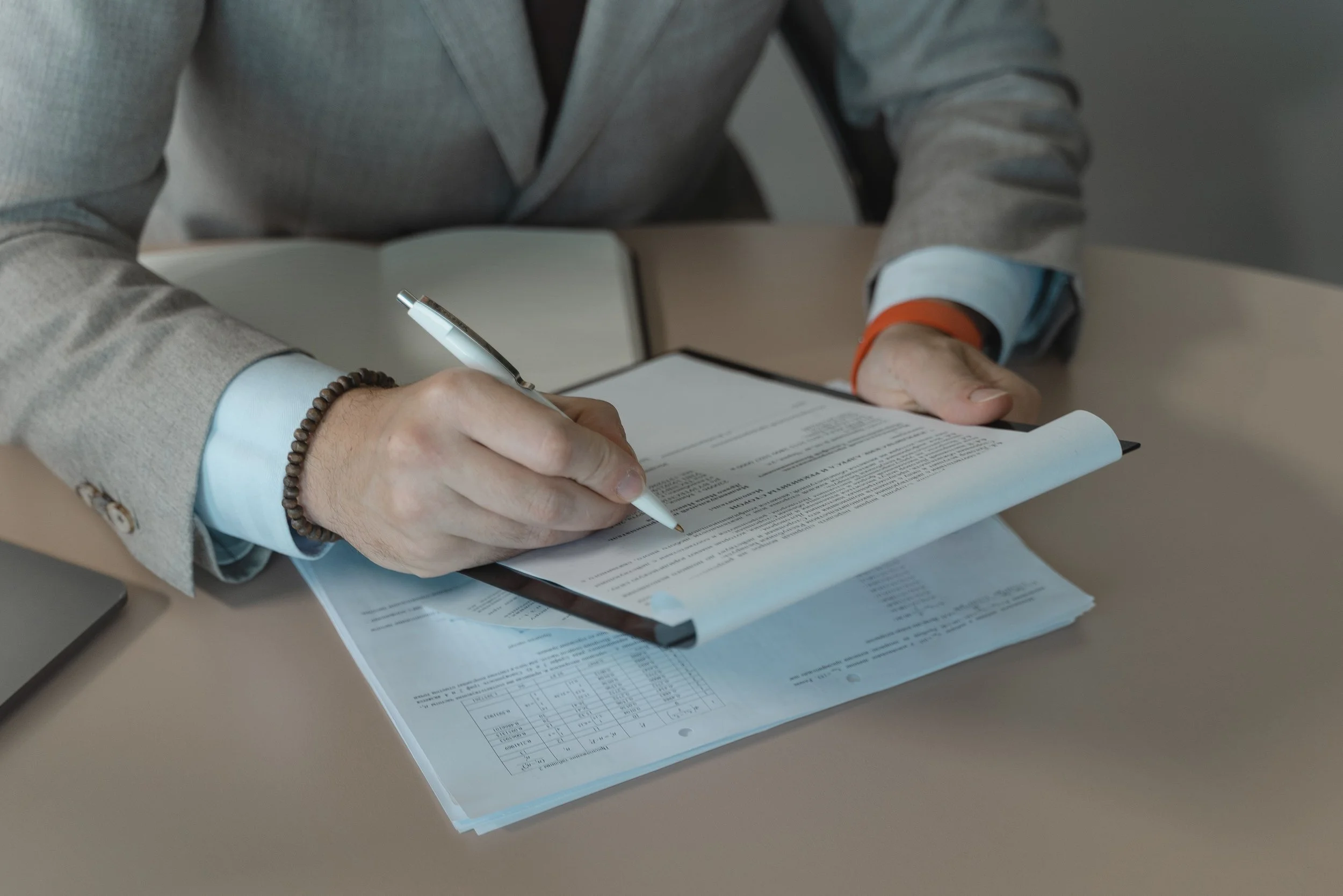 Person in a gray suit writing on a document with a white pen, seated at a table with additional papers.