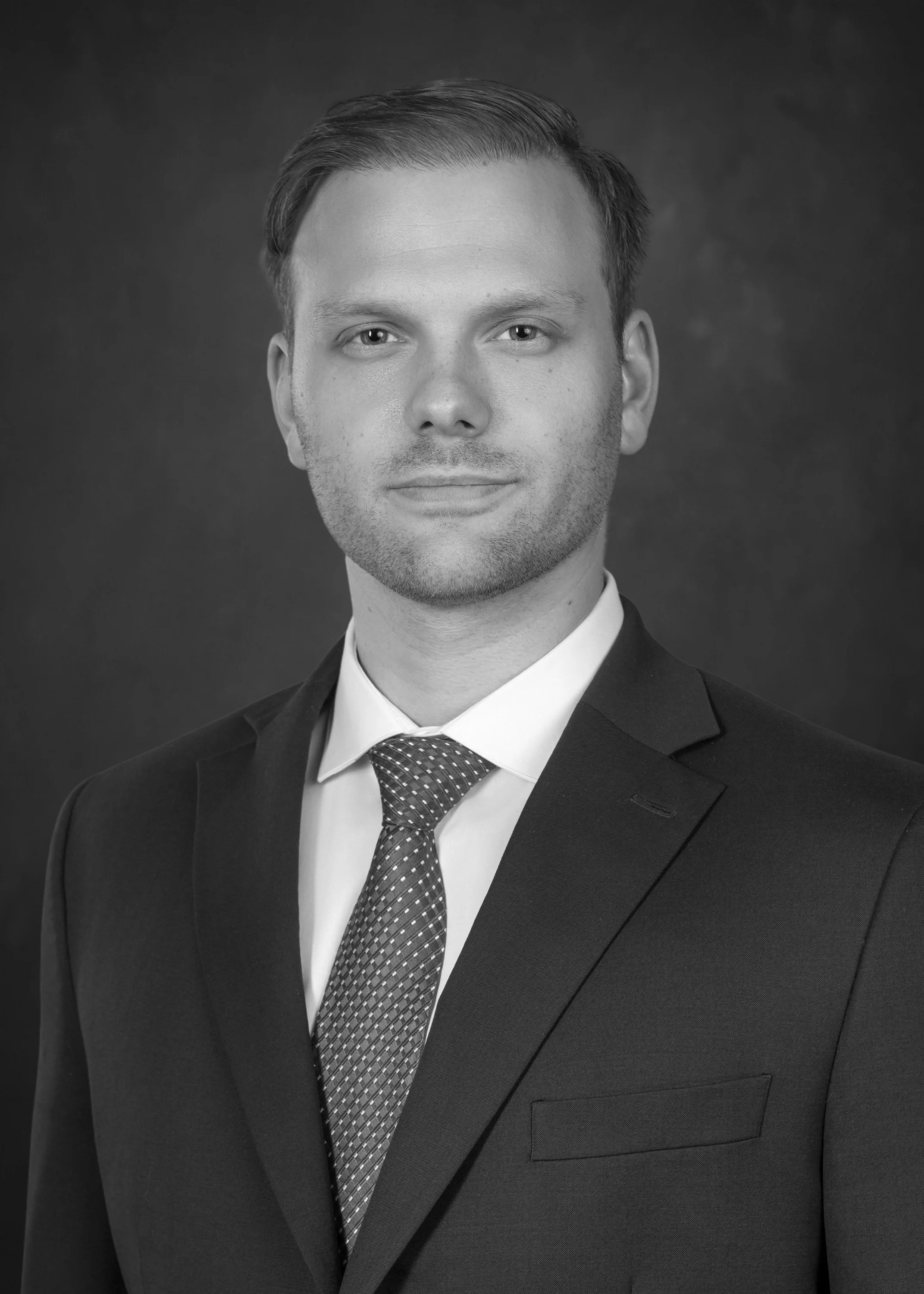 Black and white professional headshot of male attorney wearing a suit, tie, and collared shirt, against a dark background.