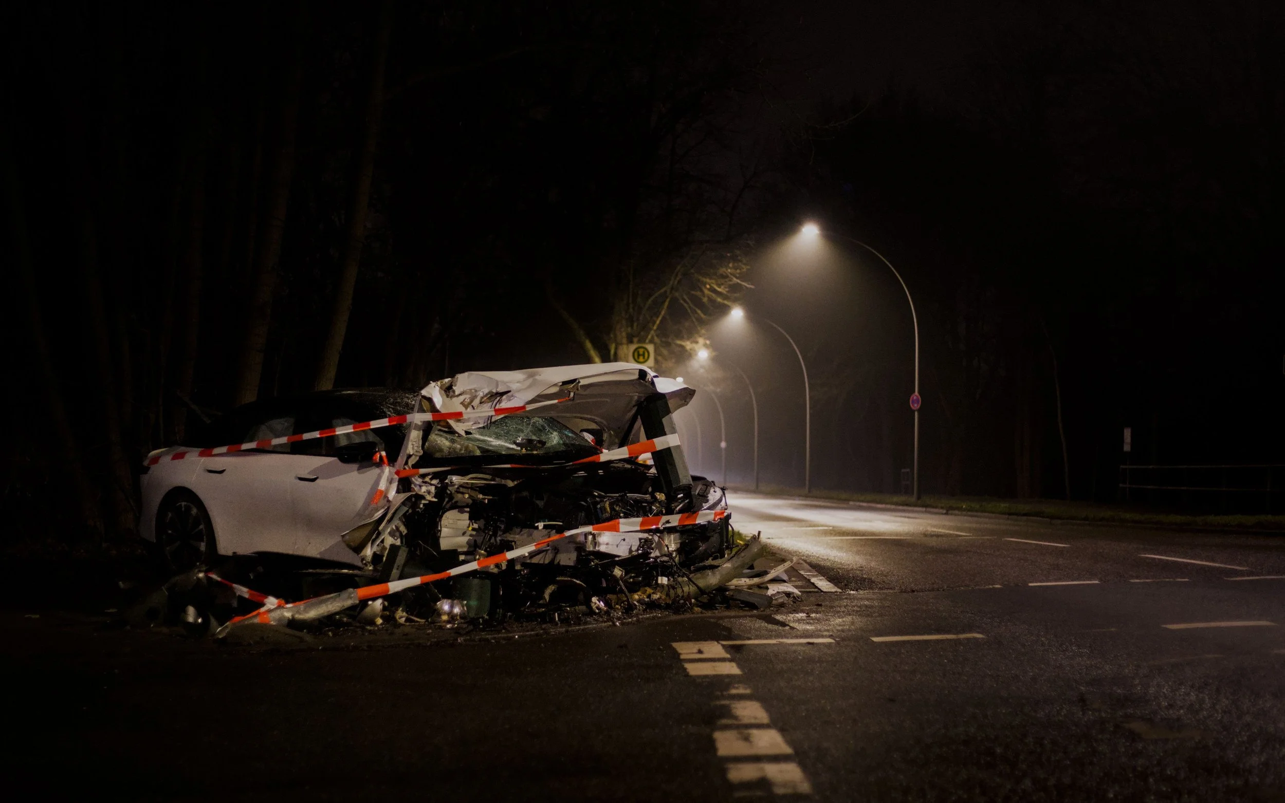 A white car with extensive front-end damage and crushed roof, surrounded by orange and white barricade tape, on a dark street at night, with streetlights in the background.