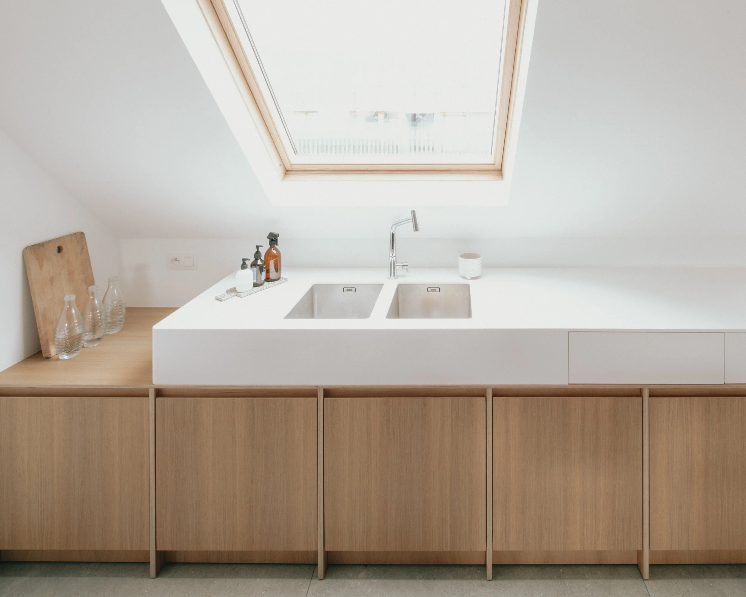 Vue d'une cuisine avec un plafond incliné, un hublot de toit, un évier double en acier inoxydable, un robinet, et des bouteilles en verre et des contenants de savon sur le plan de travail en blanc. Un panneau en bois est appuyé contre le mur à gauche