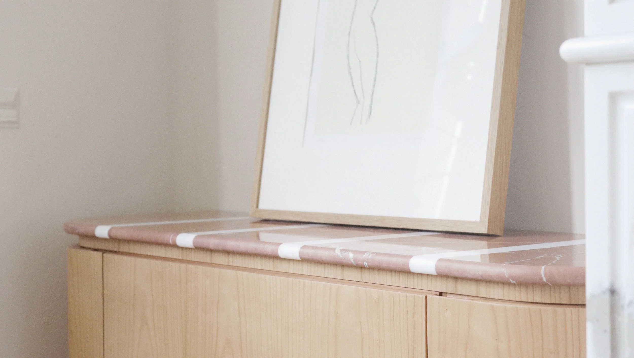 Miroir posé sur une commode en bois clair avec un profil en marbre rose, dans une pièce aux murs blancs.