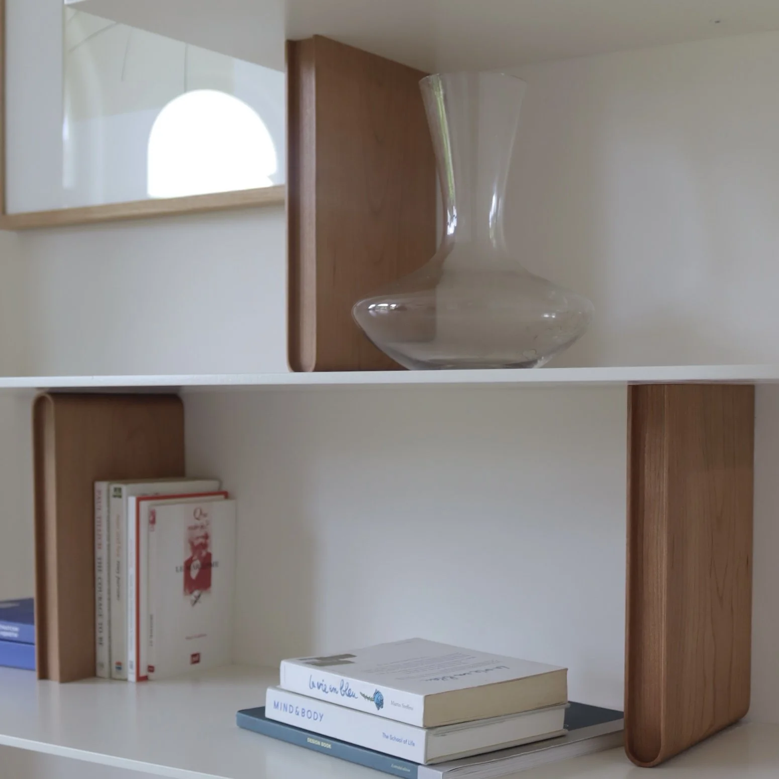 Une étagère blanche avec un livre vert et blanc, un vase en verre transparent, et une collection de livres, dont 'Mind & Body', 'The School of Life', et 'Consciousness'.