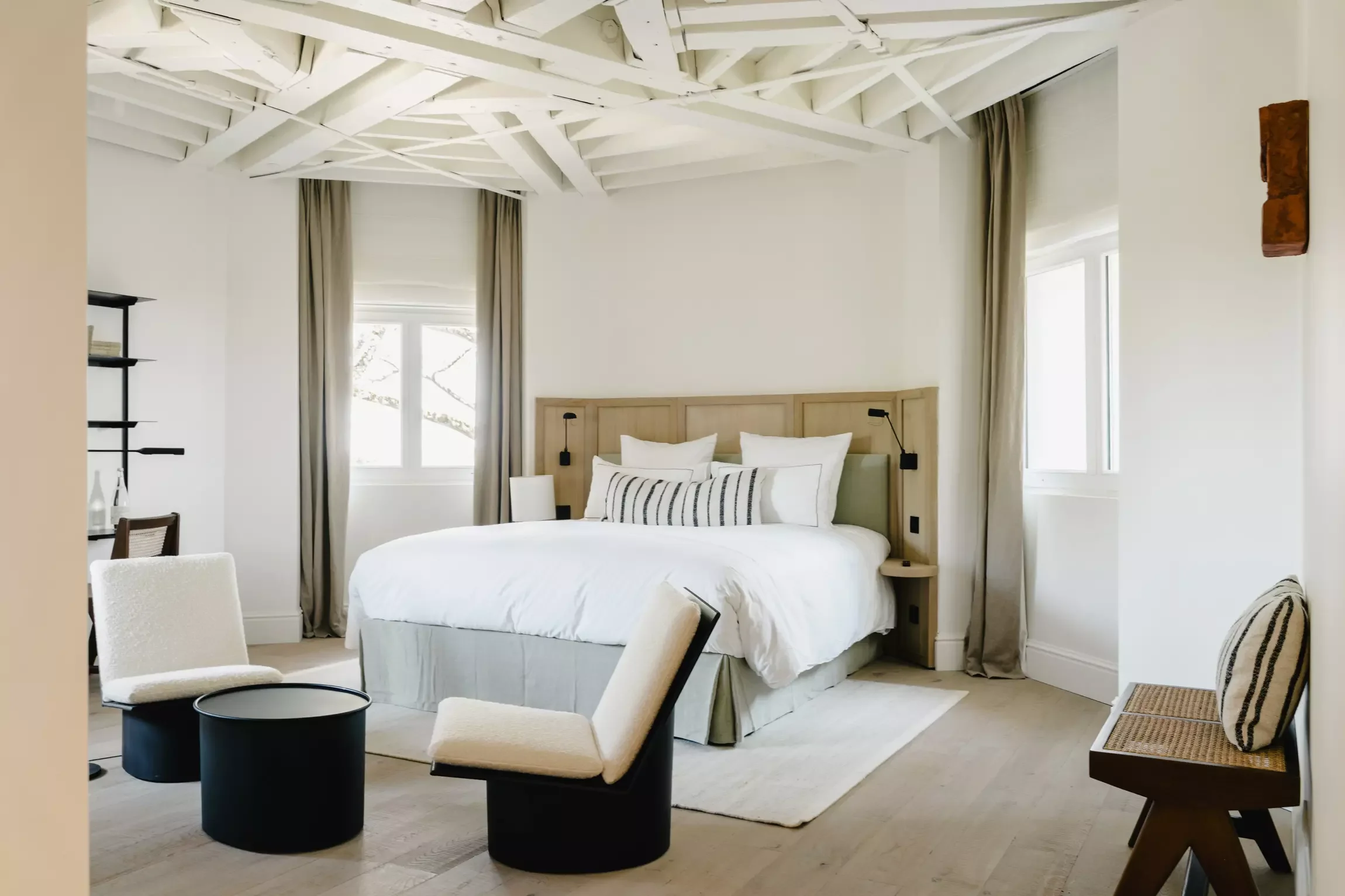 Chambre spacieuse avec un lit blanc, plusieurs fenêtres avec rideaux beiges, décoration minimaliste, meubles en bois clair, quelques chaises et tables, ambiance lumineuse.