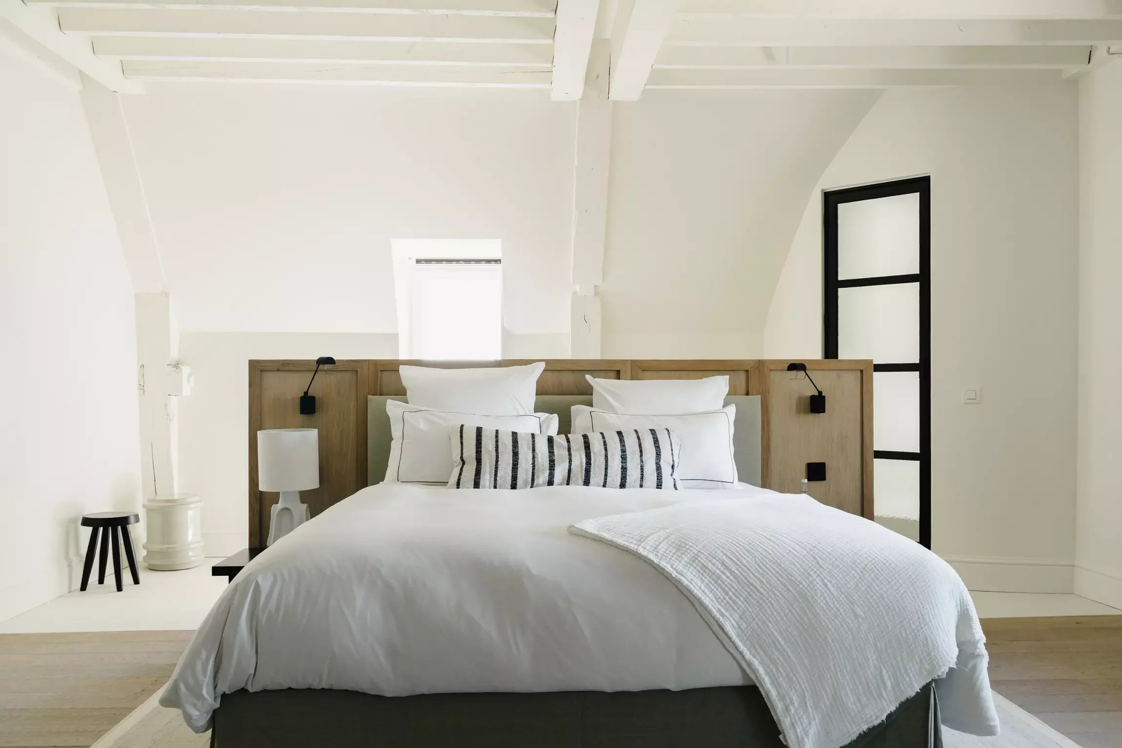 Chambre moderne avec lit fait, tête de lit en bois, deux lampes de lecture noires, une petite table noire, des coussins noirs et blancs, mur blanc avec fenêtre haute et porte en verre avec cadre noir.