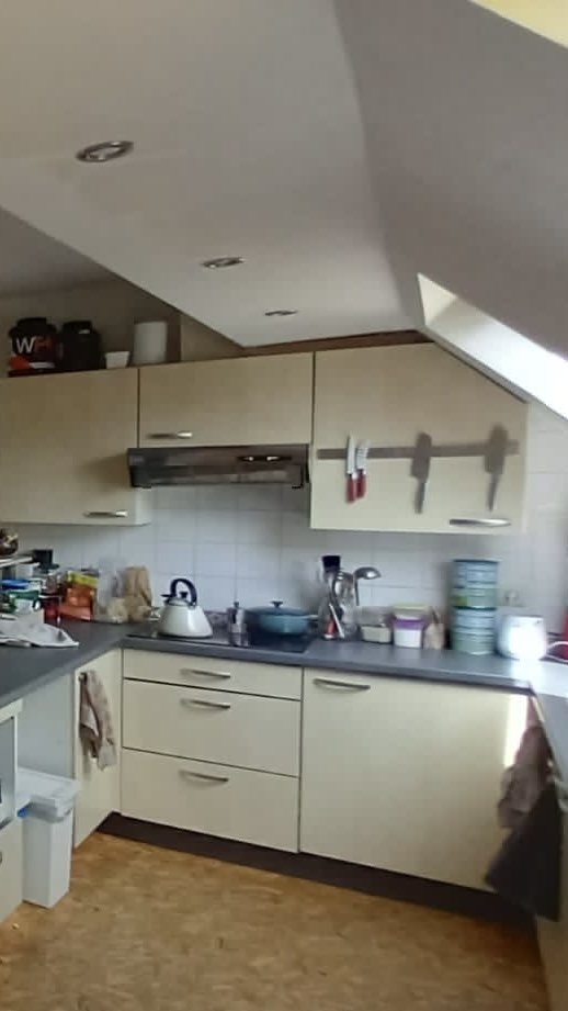 Cuisine avec des armoires beiges, un plan de travail en noir, une cuisinière à bois, un évier avec un robinet, et des objets variés sur le comptoir, sous un plafond incliné avec des spots lumineux.
