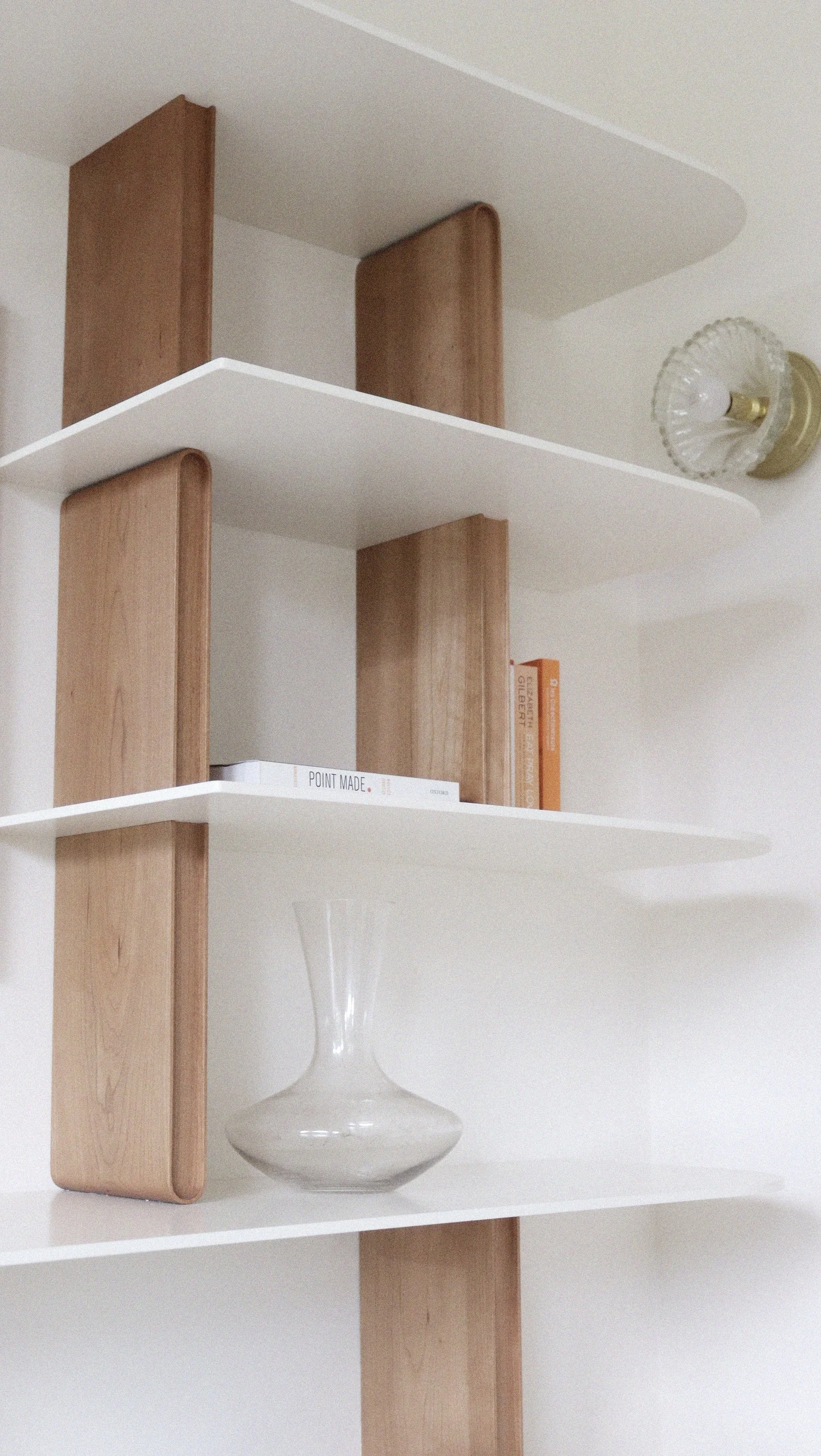 Une étagère murale blanche avec des côtés en bois, contenant un livre blanc intitulé 'POINT MADE', un livre orange, une bouteille en verre et un vase blanc.