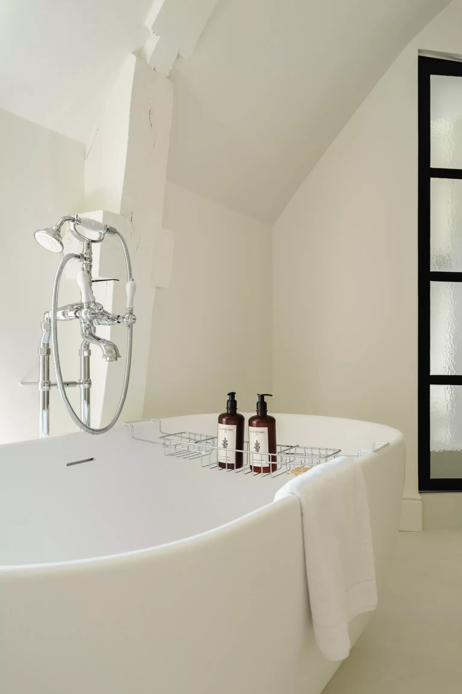 Baignoire blanche avec panier de rangement contenant deux bouteilles de savon ou shampooing, en face d'une cabine de douche avec robinetterie vintage, dans une salle de bains moderne.