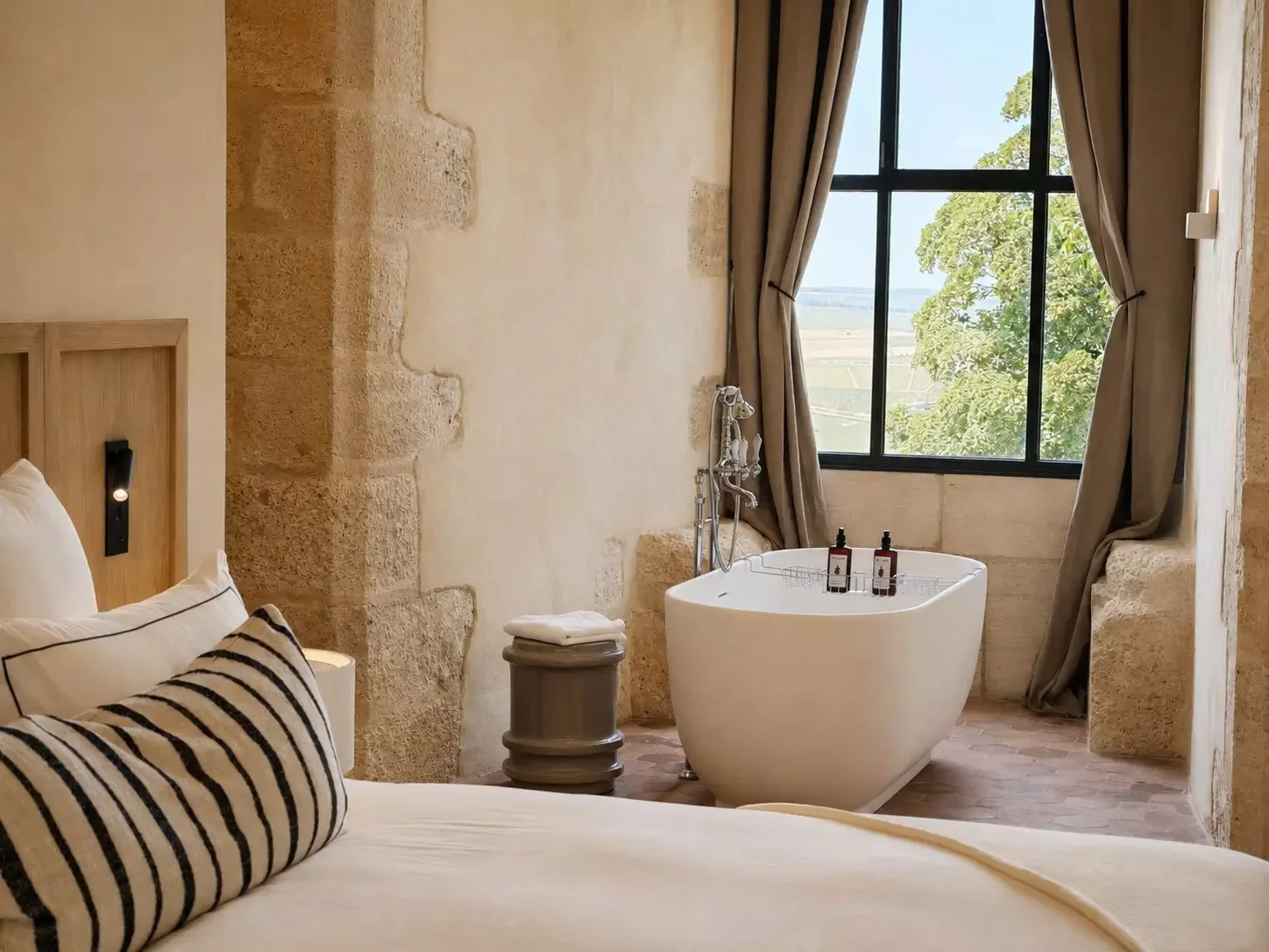 Chambre de hotel avec vue sur un paysage rural, grande baignoire à côté de la fenêtre, lit avec oreillers à rayures, murs en pierre beige, rideaux ouverts laissant entrer la lumière naturelle.