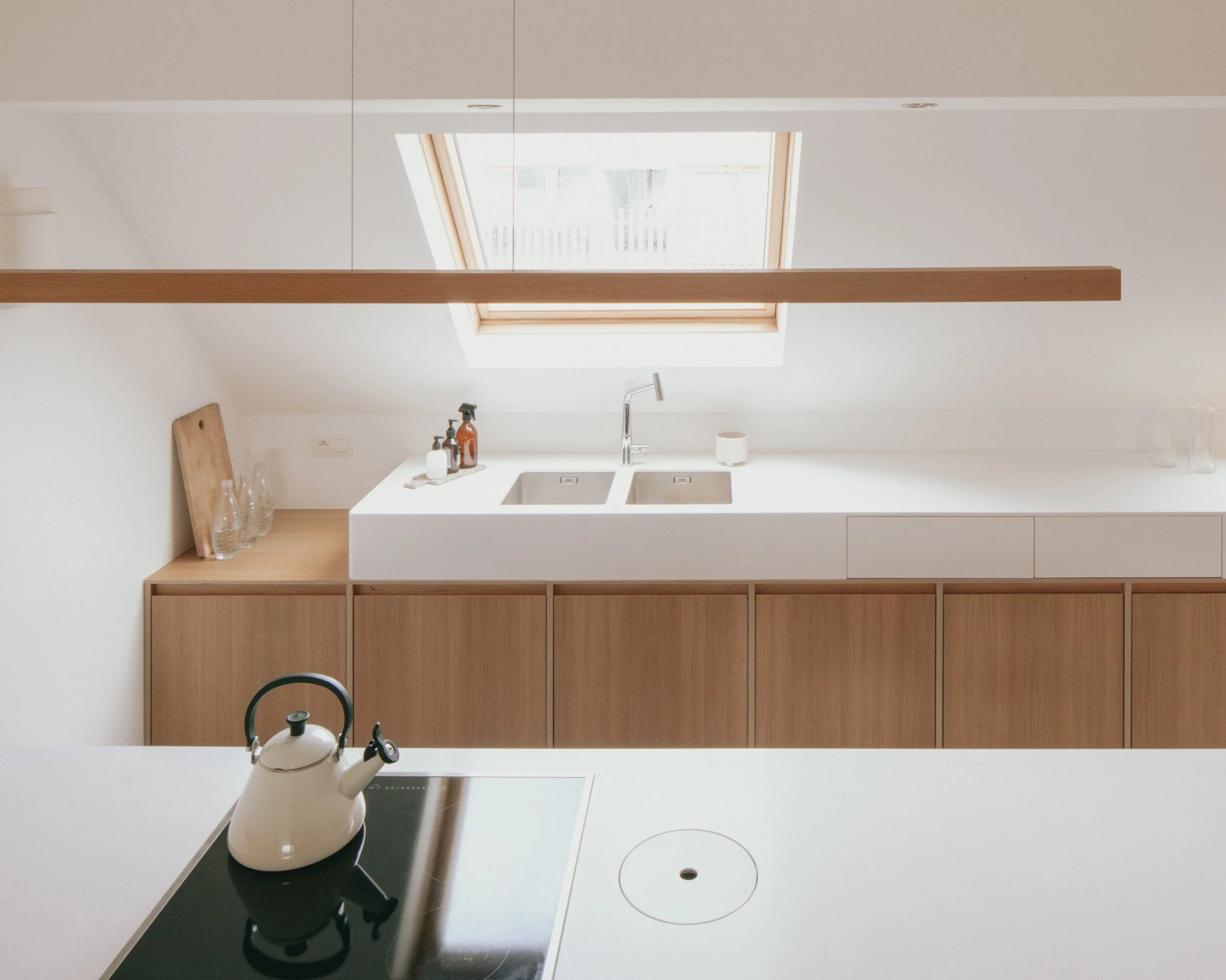 Cuisine moderne avec un évier à double bassine, plan de travail en bois clair, trousseau de vaisselle, une cafetière blanche, et un plafond avec un toit en lucarne laissant passer la lumière naturelle.