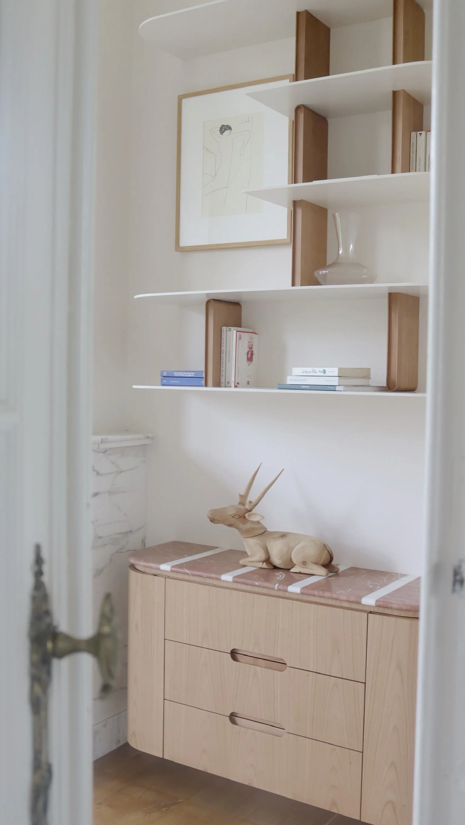 Une étagère blanche avec des livres, une assiette en verre et une illustration encadrée, au-dessus d'un meuble en bois clair avec un décor de tête de cerf en bois.
