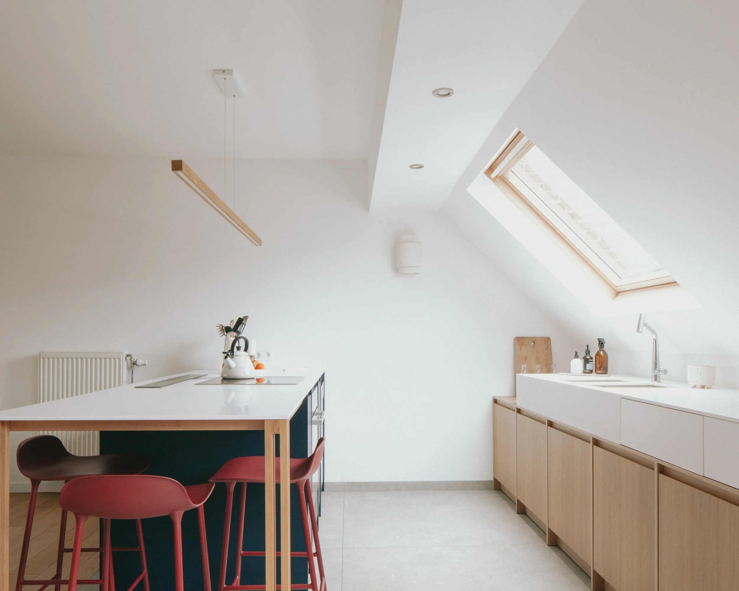 Cuisine moderne avec des meubles en bois clair, un plan de travail blanc, un évier, un plafond avec une lucarne, et des chaises rouges autour de la table.