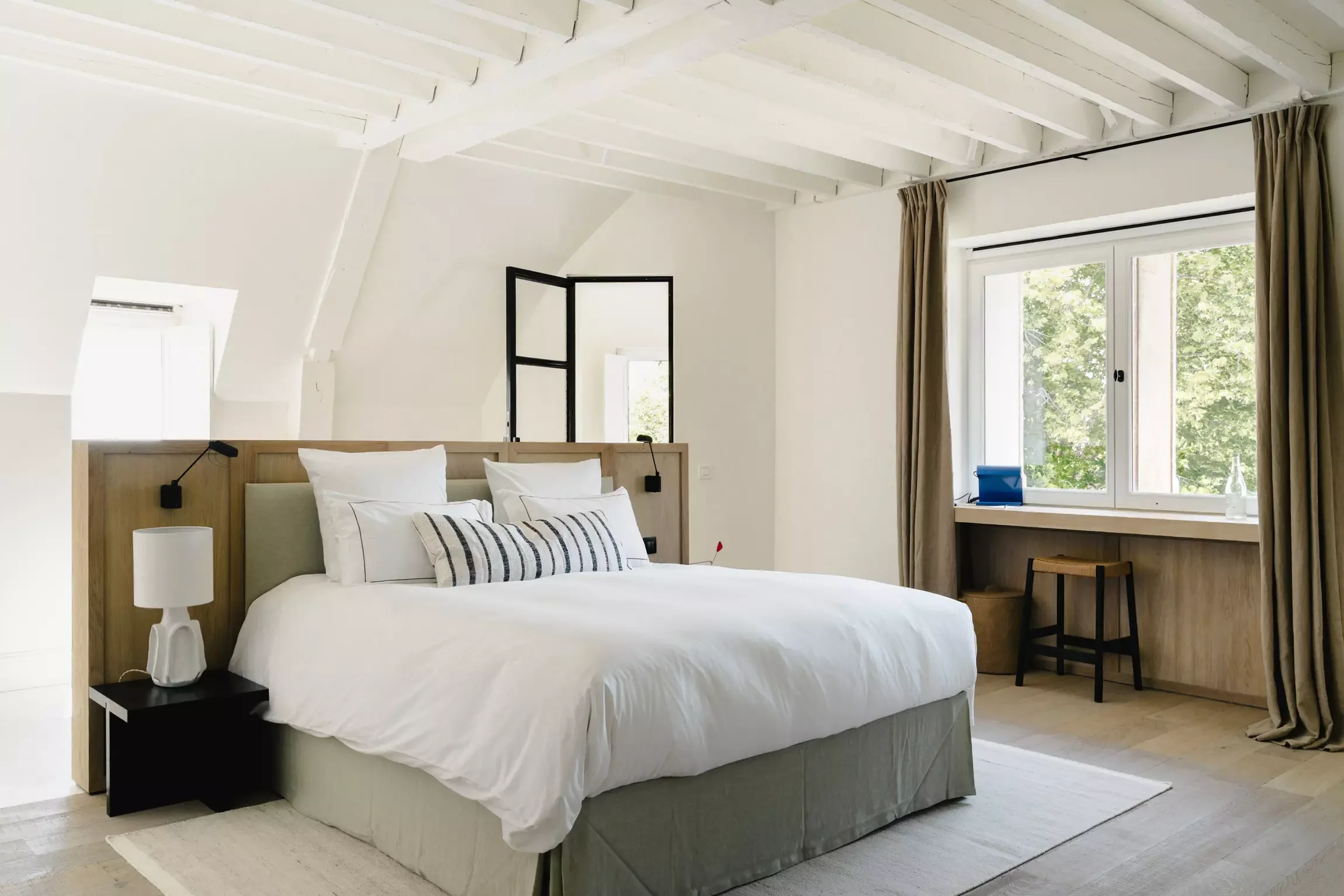 Chambre lumineuse avec un lit blanc, deux lampes de chevet noires, grandes fenêtres laissant entrer la lumière naturelle, et une décoration neutre avec des rideaux beiges.