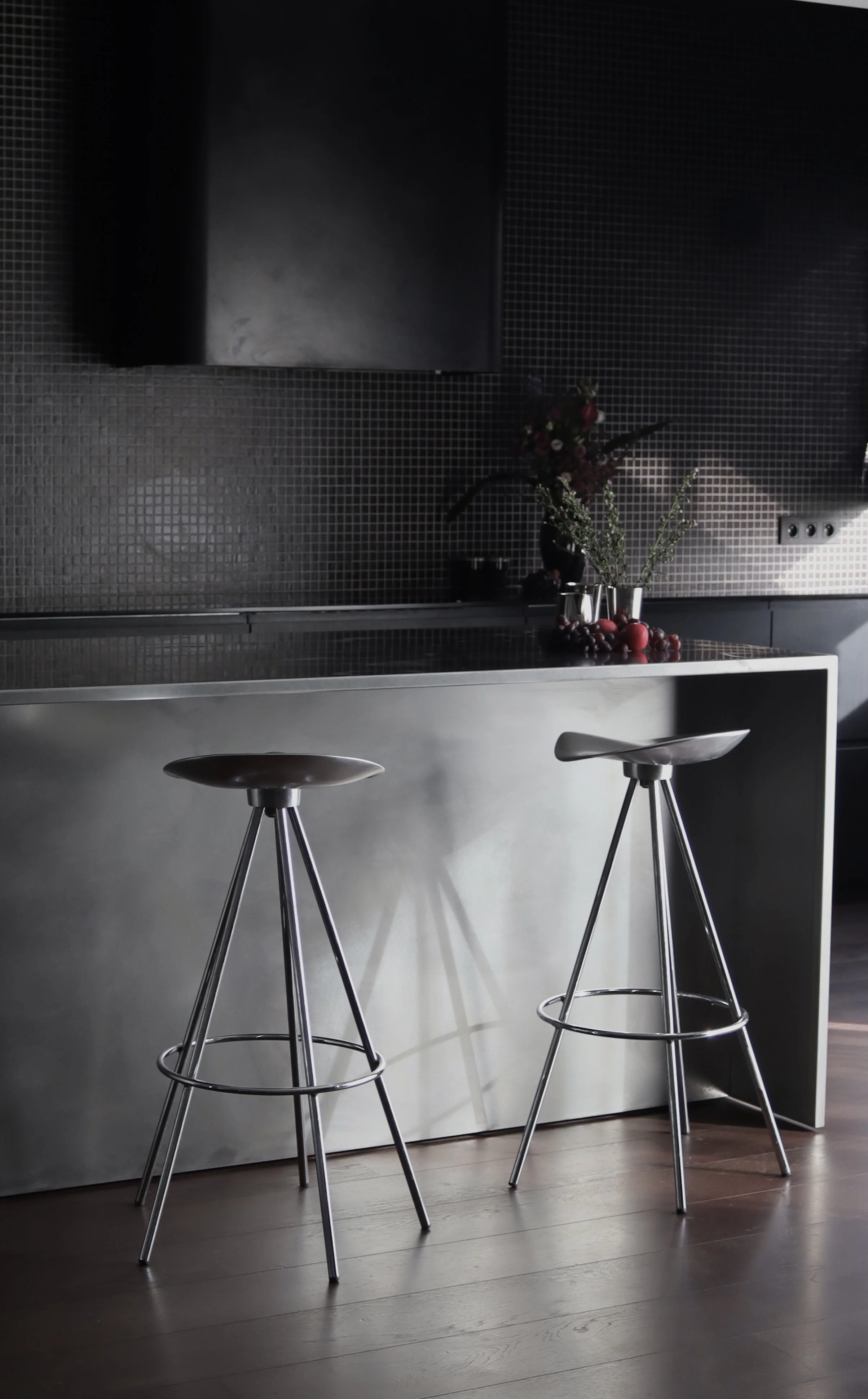 Cuisine moderne avec deux chaises hautes en métal au premier plan, un comptoir en acier inoxydable, un vase avec des fleurs et des fruits rouges, mur noir avec carrelage en mosaïque, et une prise électrique à droite.