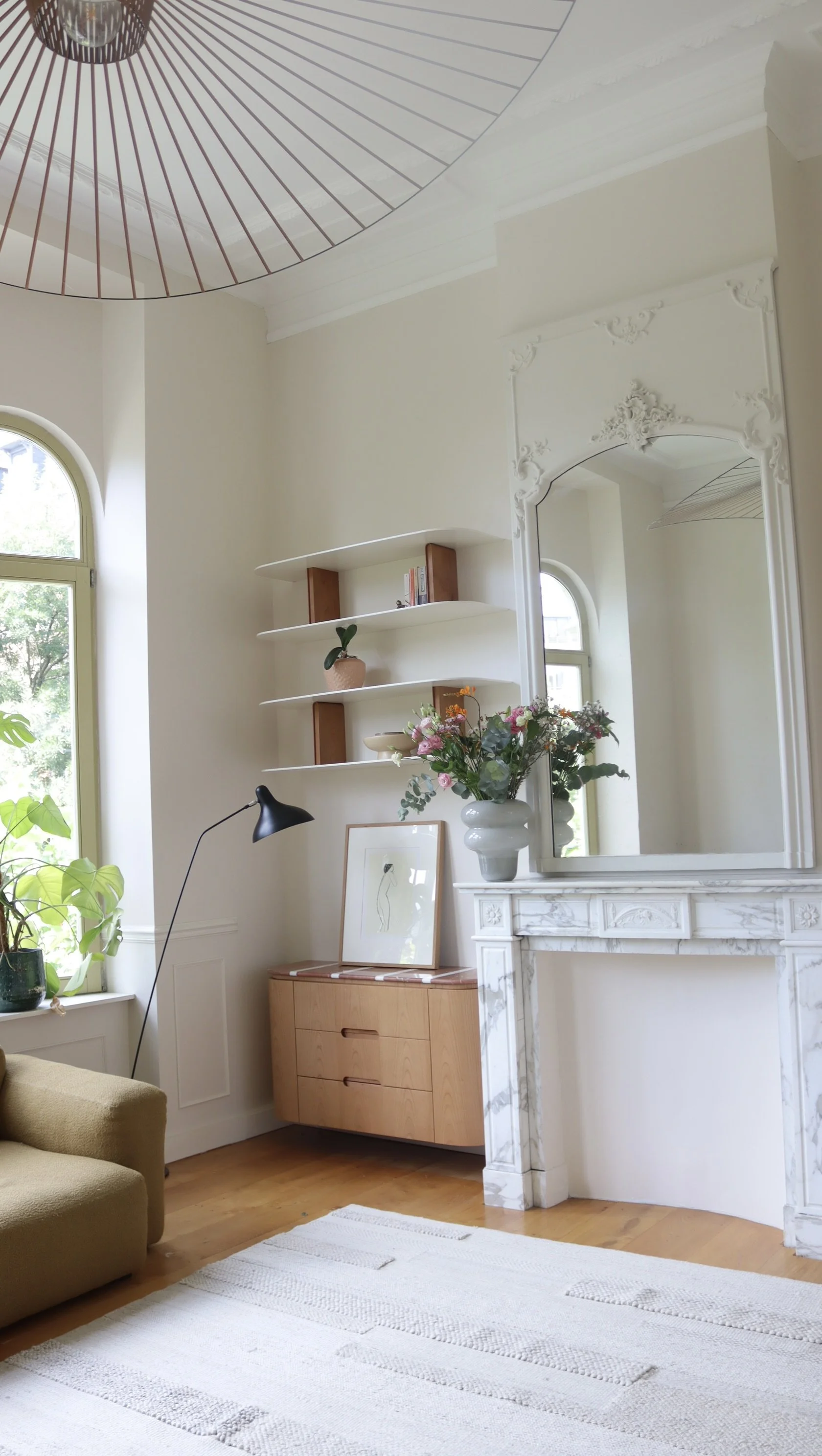 Intérieur d'un salon avec une cheminée en marbre blanc, une grande fenêtre en arc, une étagère blanche avec des livres et une plante, un miroir ancien, un vase avec des fleurs, un bureau en bois clair, un lampadaire noir, un tapis blanc à rayures et 