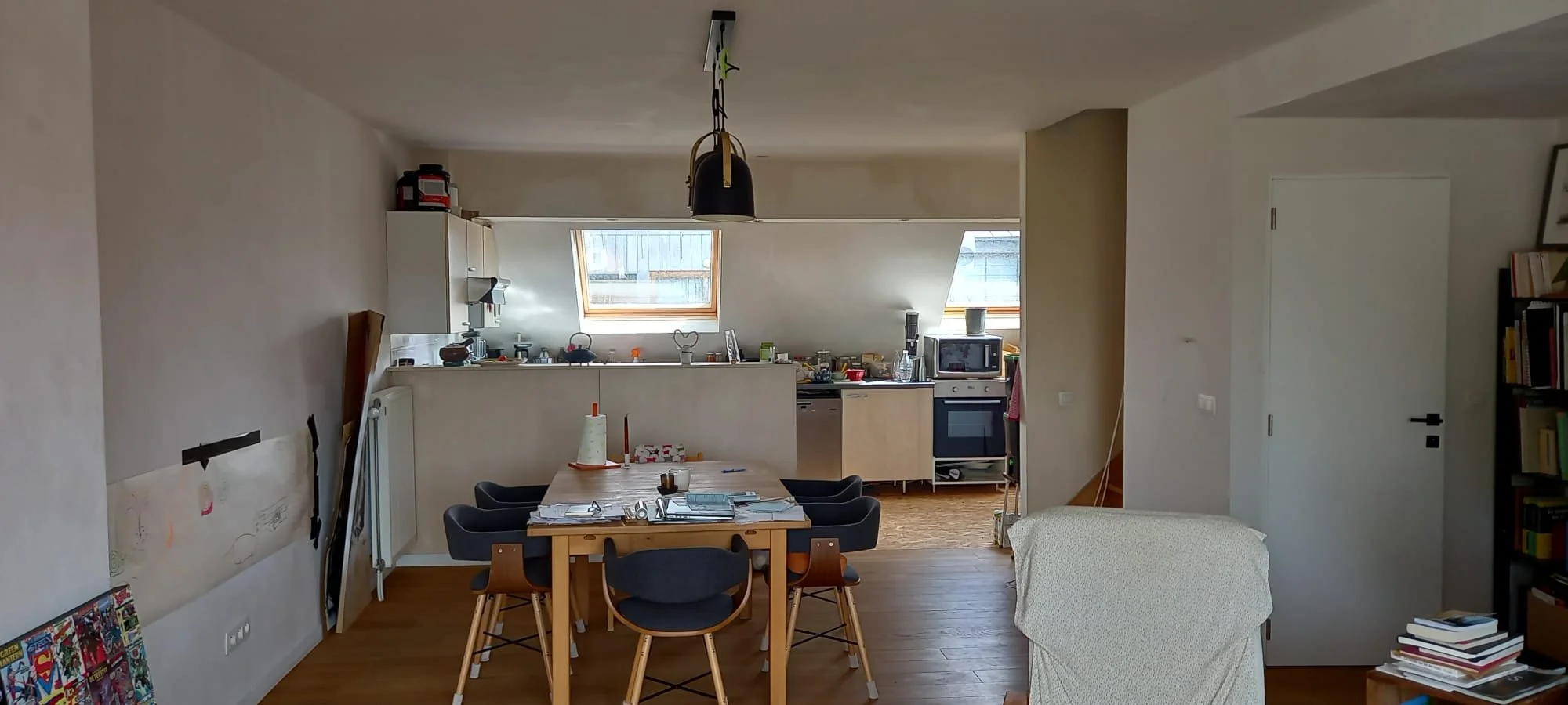 Intérieur d'une cuisine et salle à manger avec deux fenêtres en toiture, un tableau avec des comics en bas à gauche, une table en bois avec cinq chaises, un sofa blanc en premier plan, une étagère remplie de livres à droite, et un mur blanc. Éléments de décoration et objets de cuisine sur la table et le plan de travail.