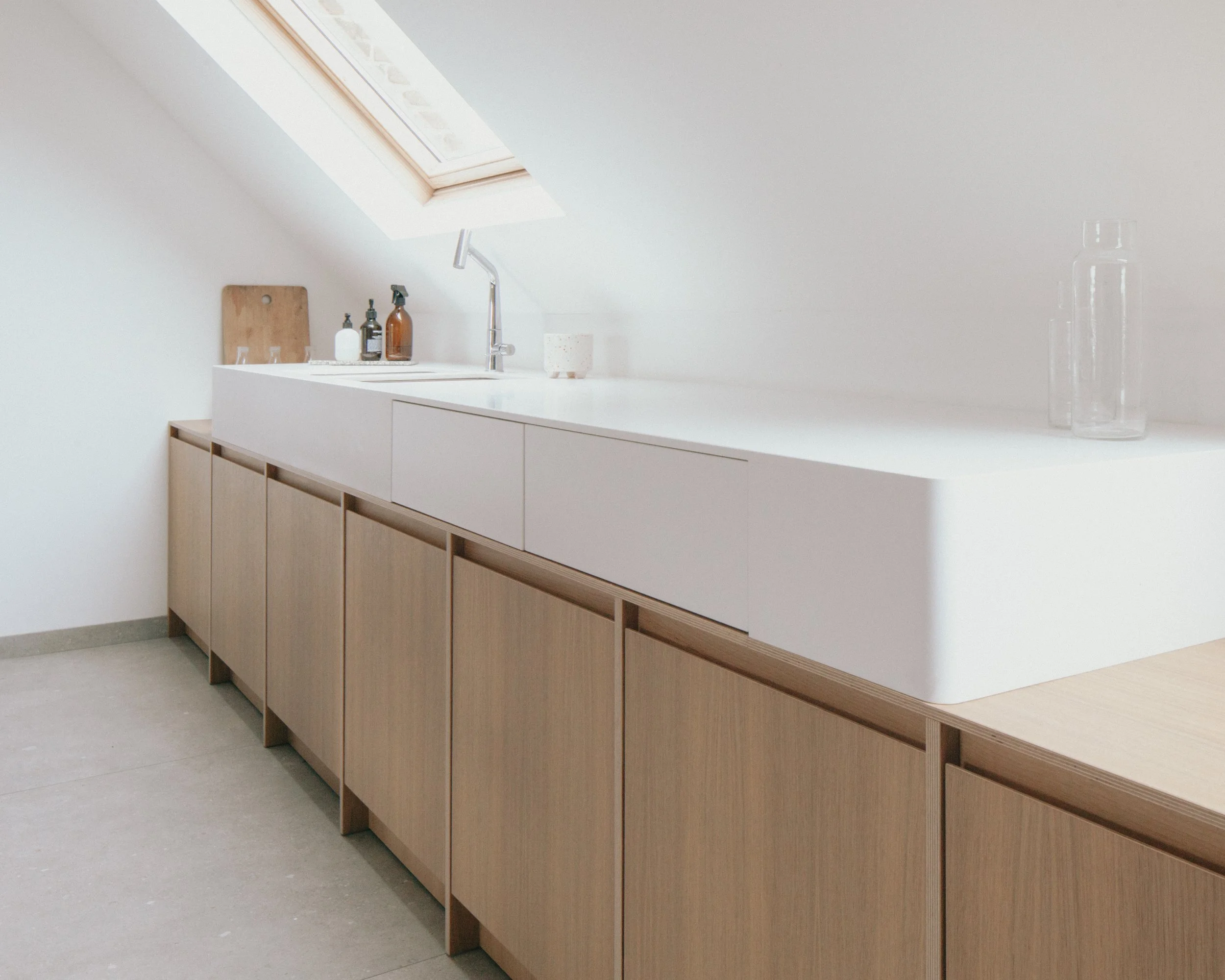 Cuisine minimaliste avec un plan de travail blanc, des bouteilles en verre et en plastique, et des meubles en bois clair.