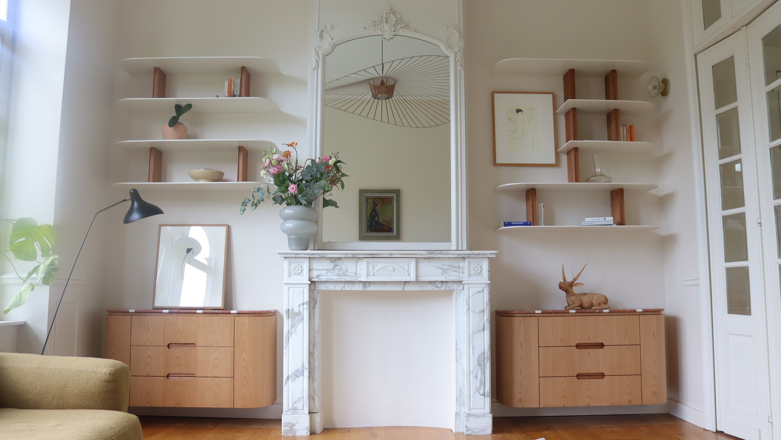 Intérieur d'un salon avec cheminée en marbre, étagères blanches avec objets décoratifs, reflet dans un grand miroir, plante verte, tableau, et décoration en bois, style moderne et épuré.